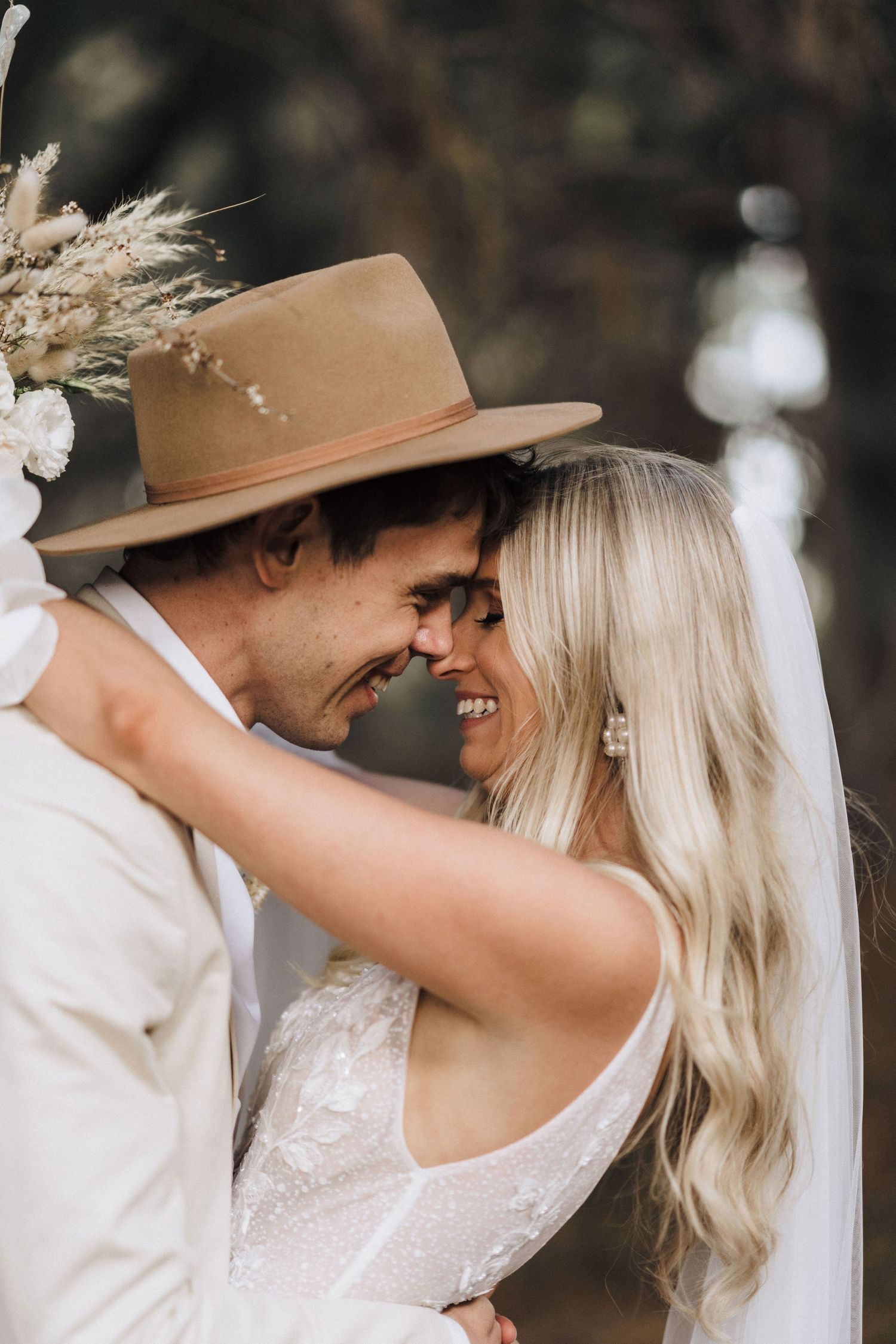 Romantic couple sharing an intimate moment outdoors under the brim of a vintage-style hat and pampas grass decor.