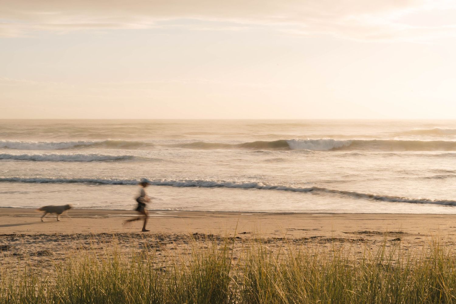 Silhouetted person jogging on misty beach at sunset with ocean waves and beach grass in foreground.