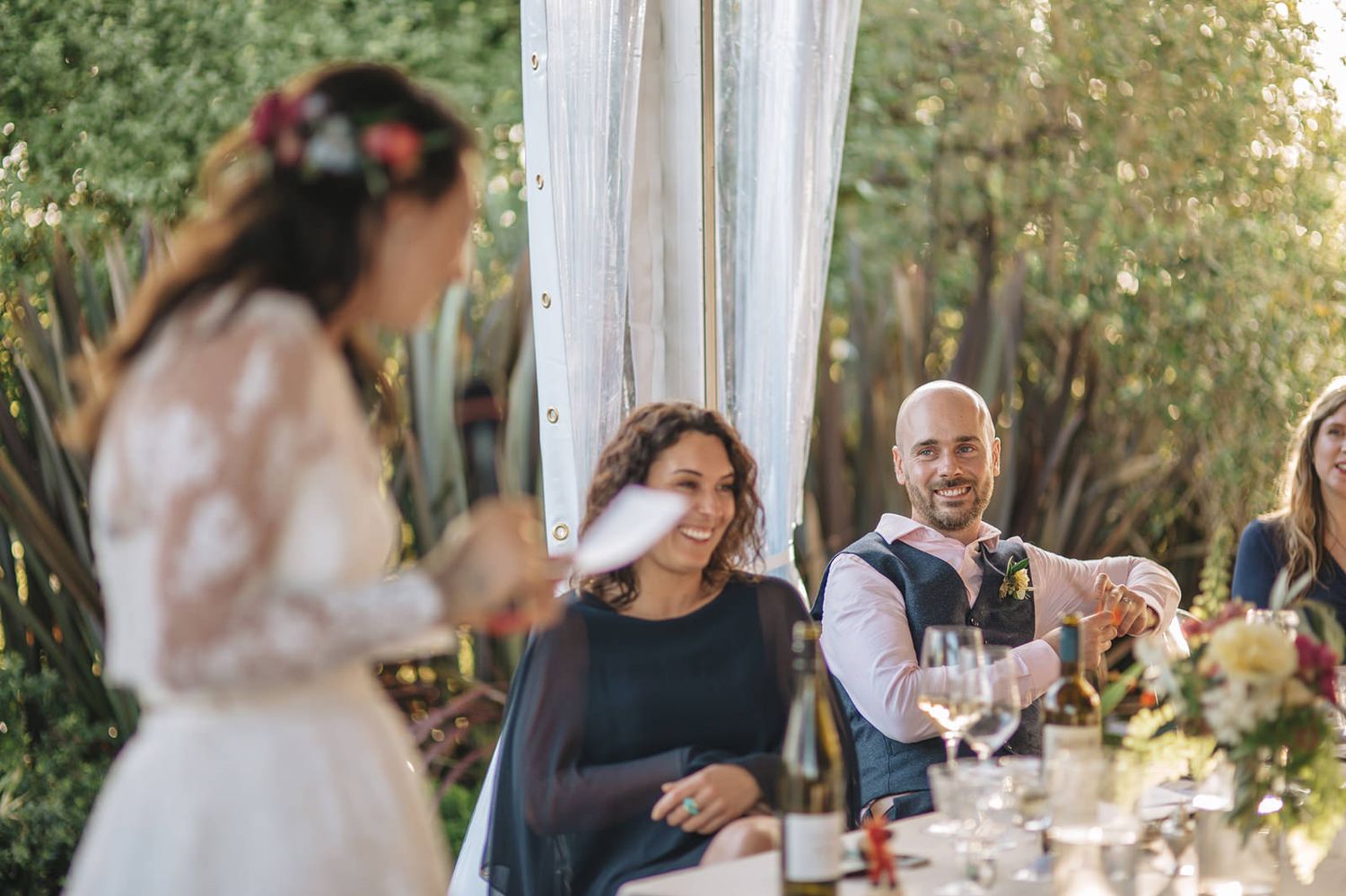 Wedding reception guests seated at long tables outdoors under string lights celebrating with wine glasses and toasts.