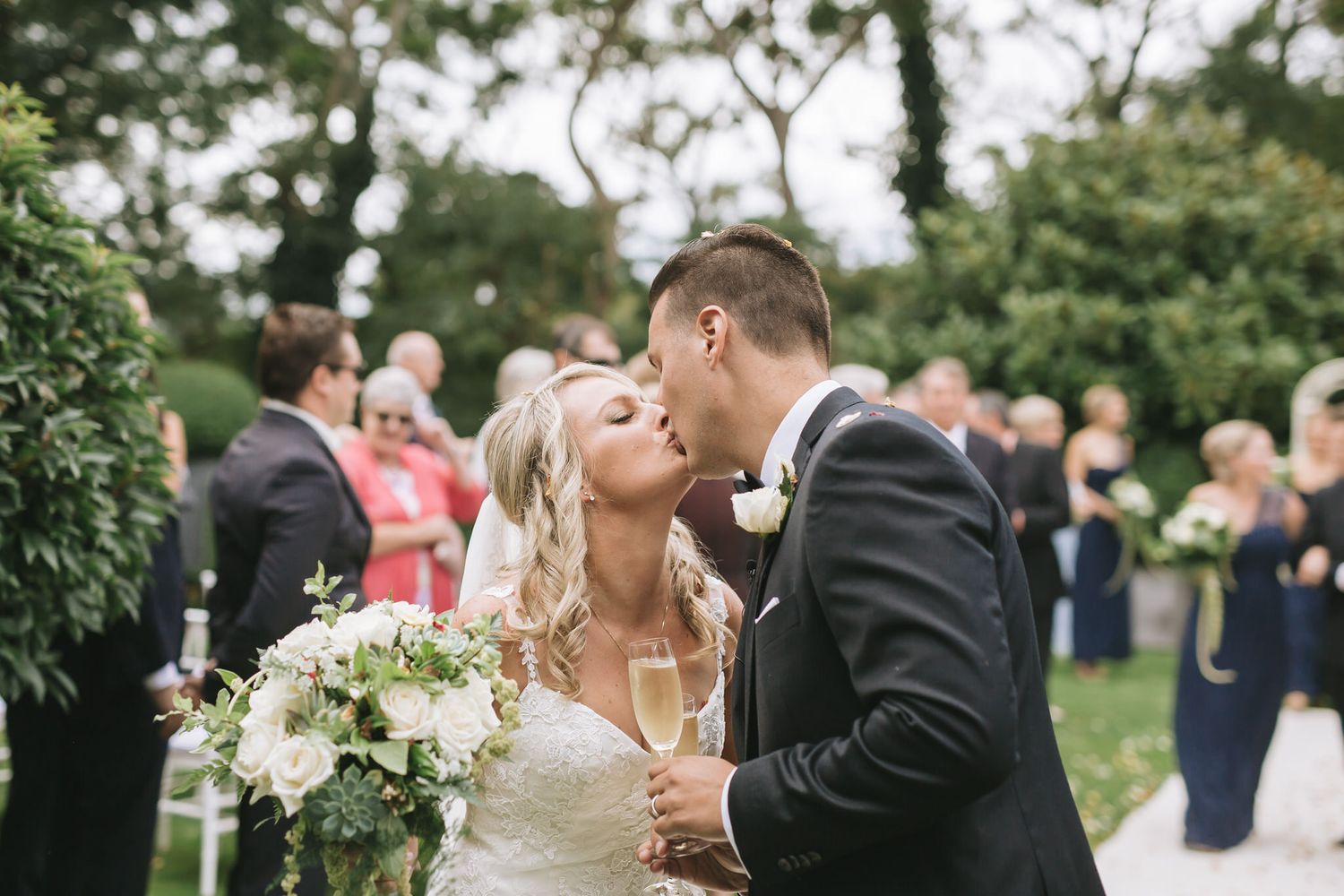 A romantic moment between newlyweds sharing a kiss in a garden setting surrounded by wedding guests.