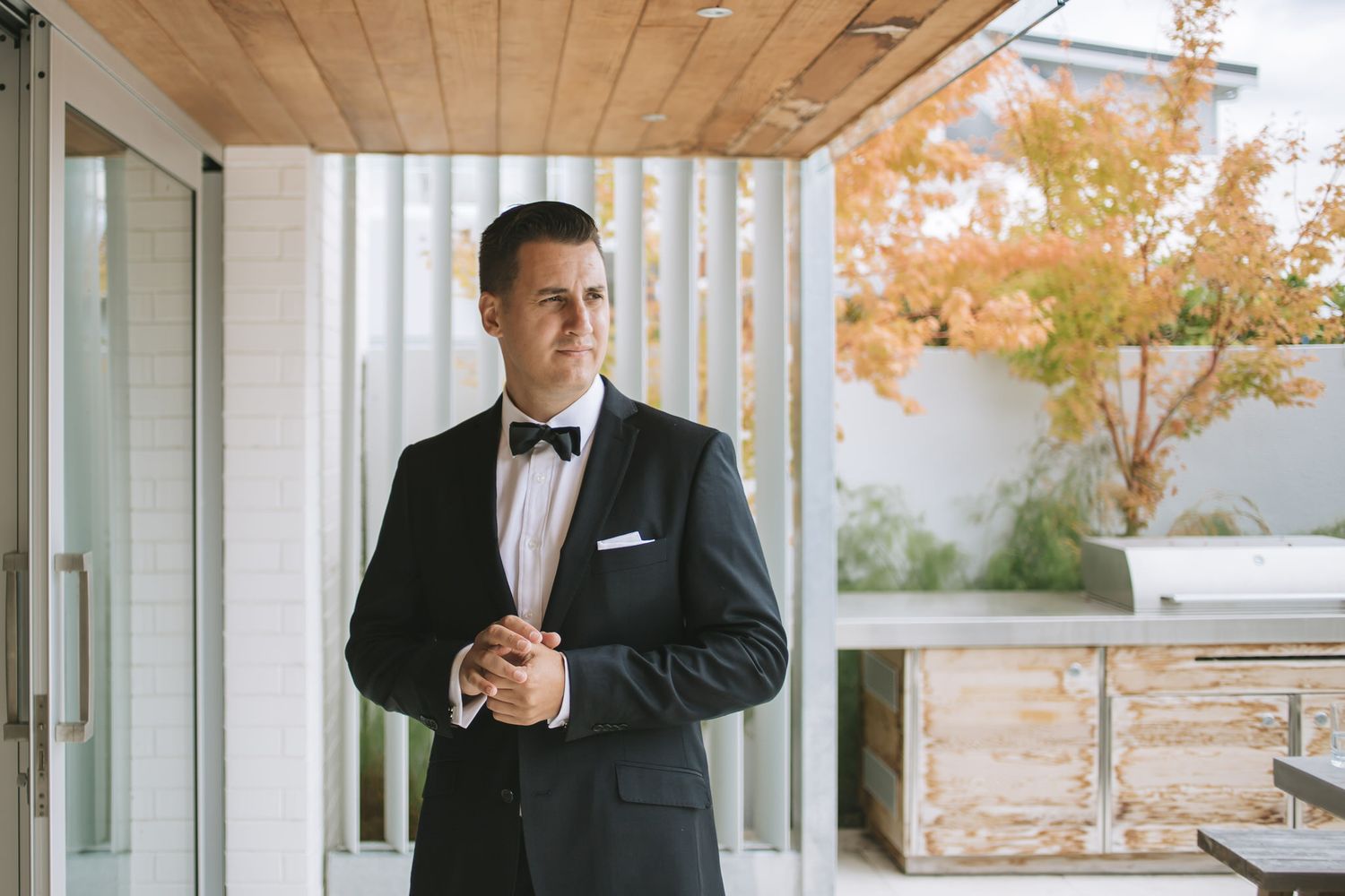 A groom in a classic black tuxedo stands in an elegant indoor venue with wood panel ceilings.