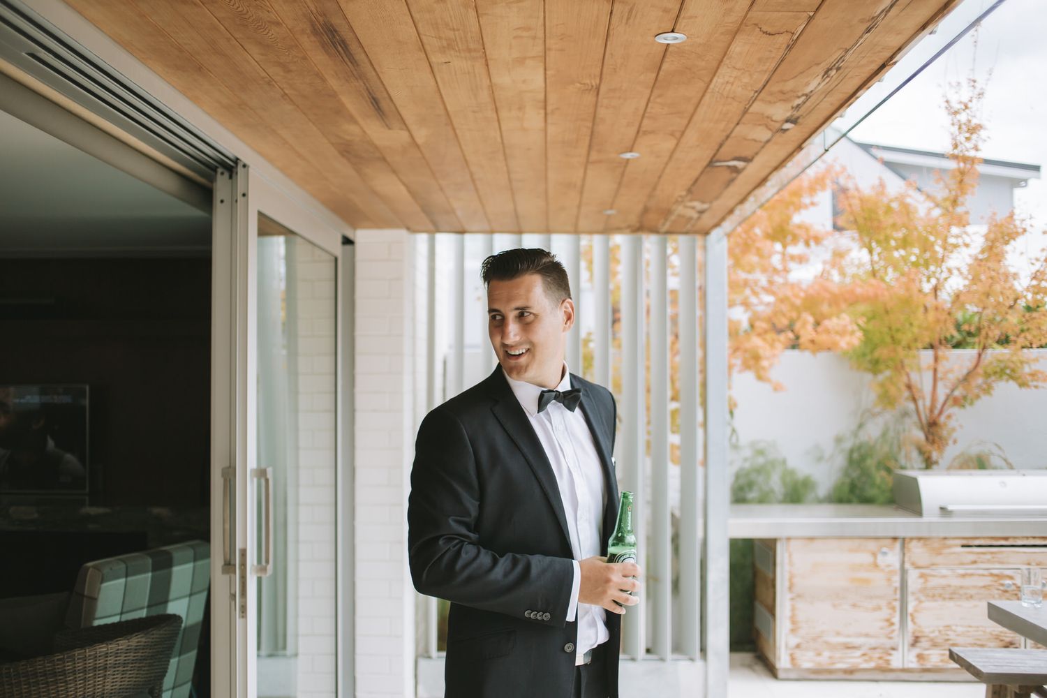 A person in a black tuxedo and bow tie stands in a covered porch area with wood paneled ceiling and fall foliage in background.