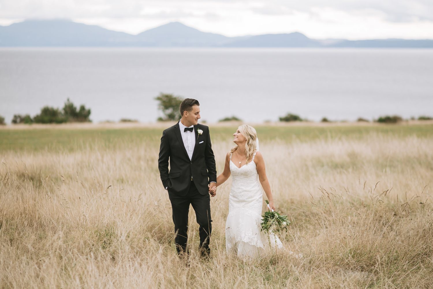 A wedding couple walks through a golden grass field with mountains and water in the background.