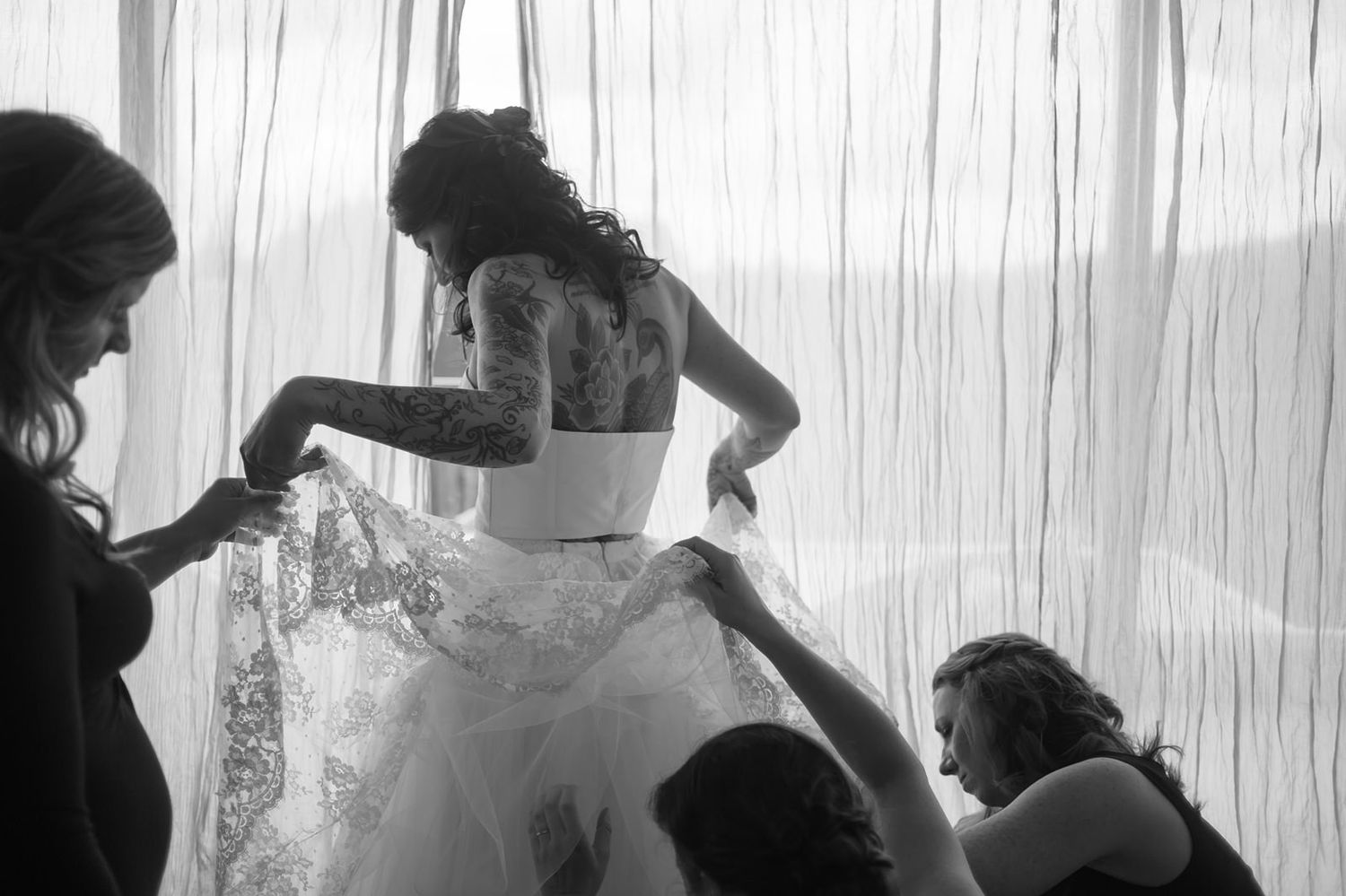 A silhouetted group helps dress someone in a wedding gown behind sheer curtains in black and white.