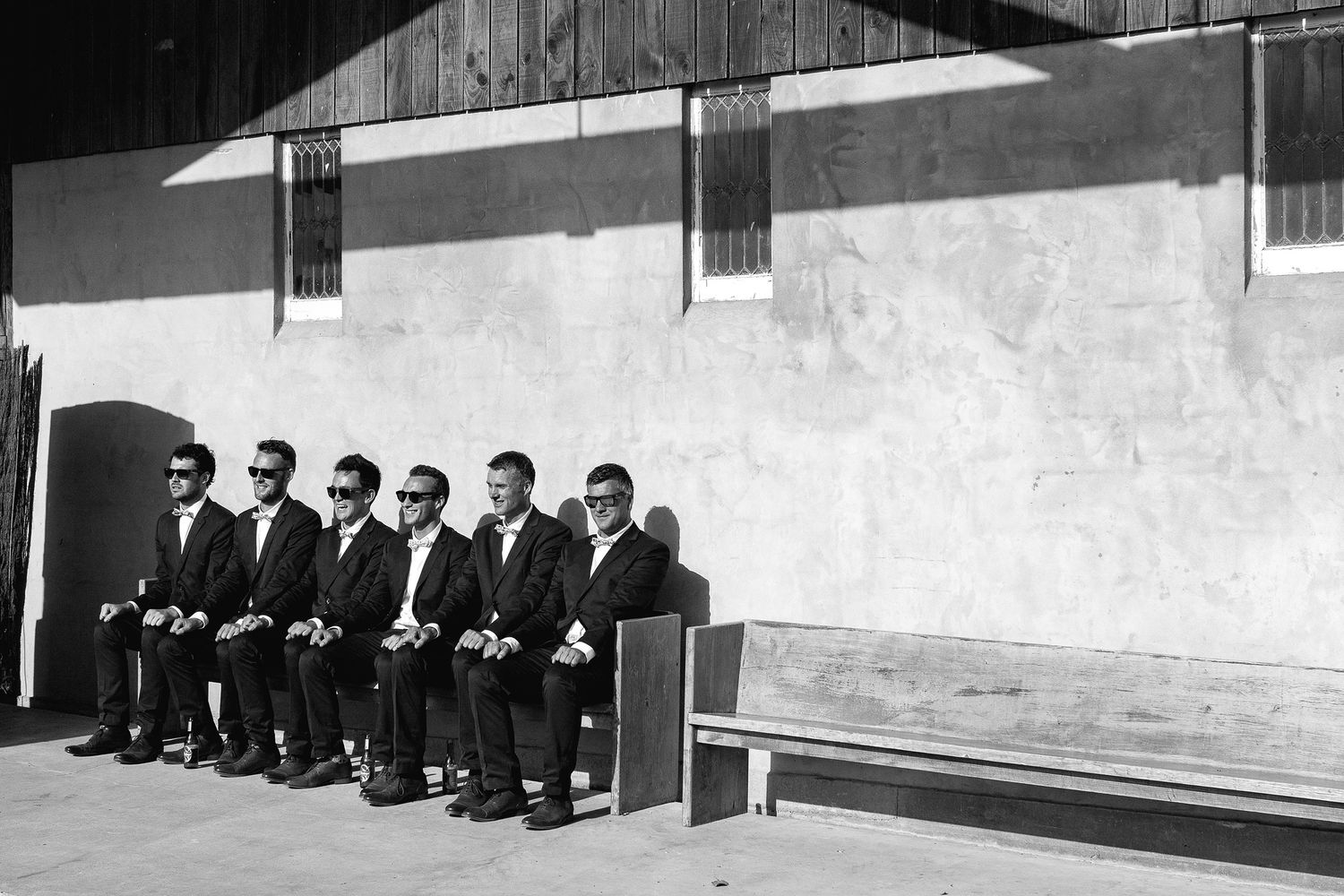 Group of men in dark suits seated on a bench against a white wall in a black and white vintage photograph.