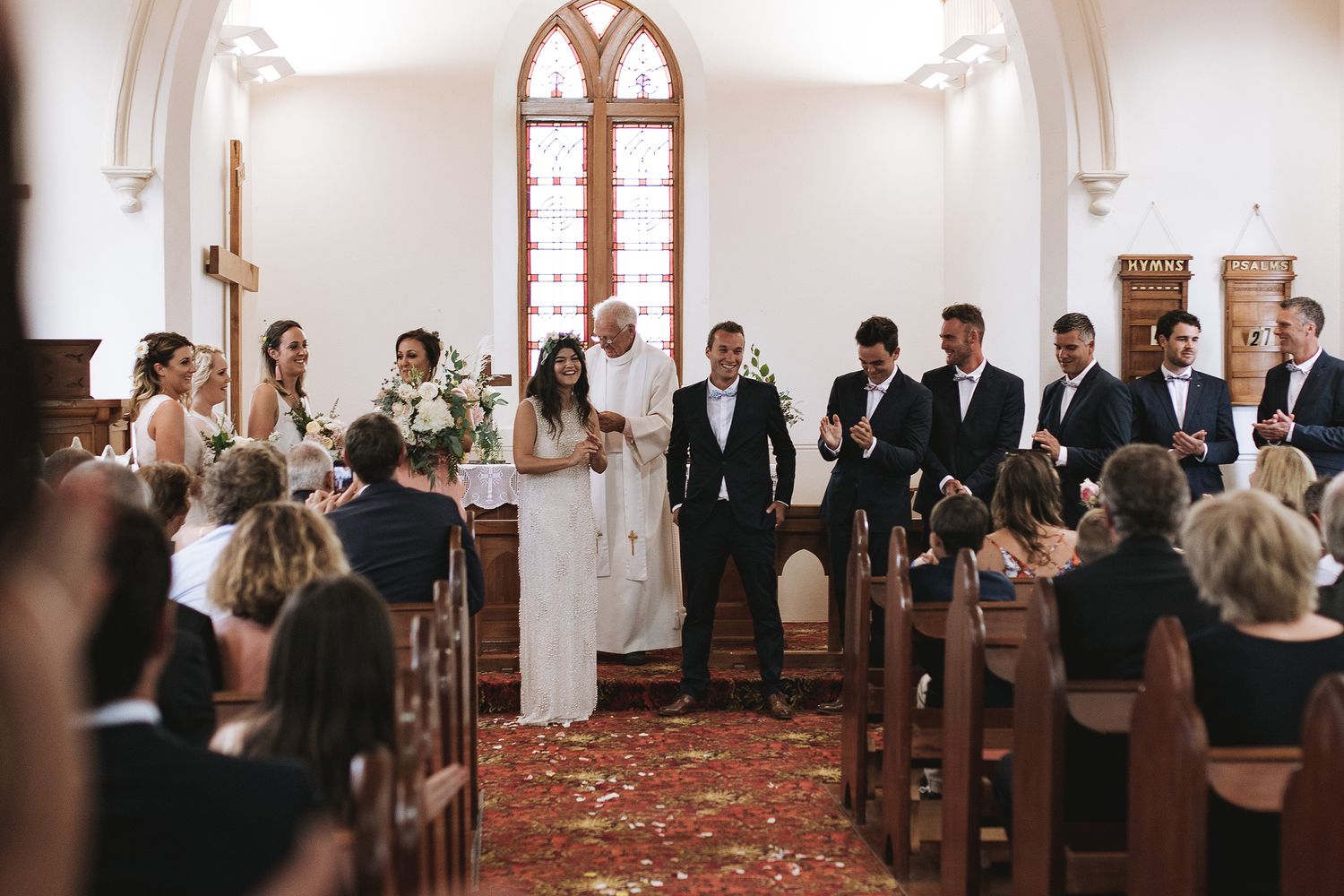 People stand in a church aisle during a traditional wedding ceremony while a couple exchanges vows at the altar.