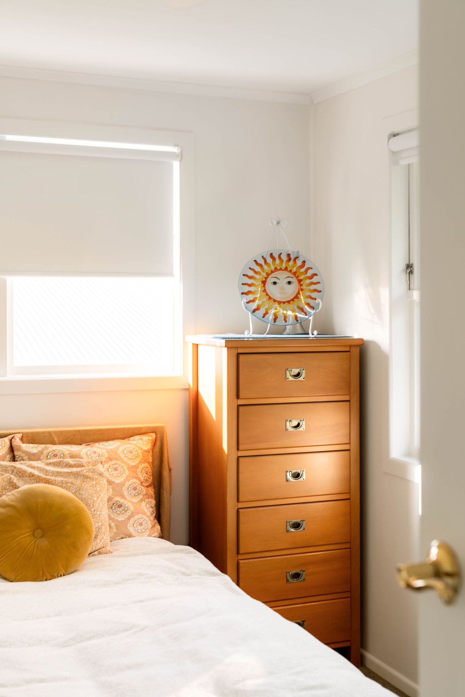 White bedding and wooden dressers create a bright, minimal bedroom with sunlit windows and decorative sun wall art.
