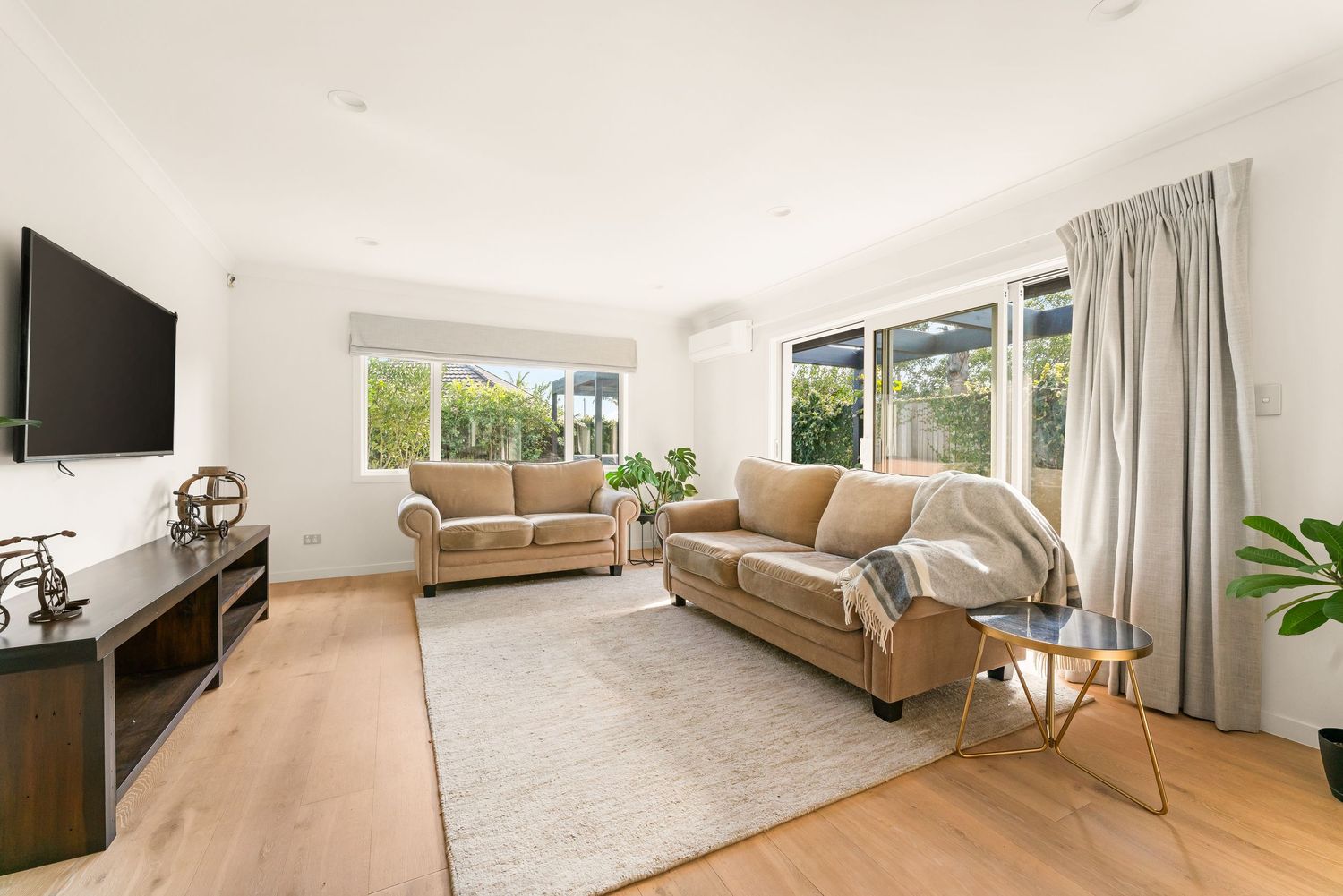 Modern living room with tan leather couches, wall-mounted TV, and hardwood flooring bathed in natural light.