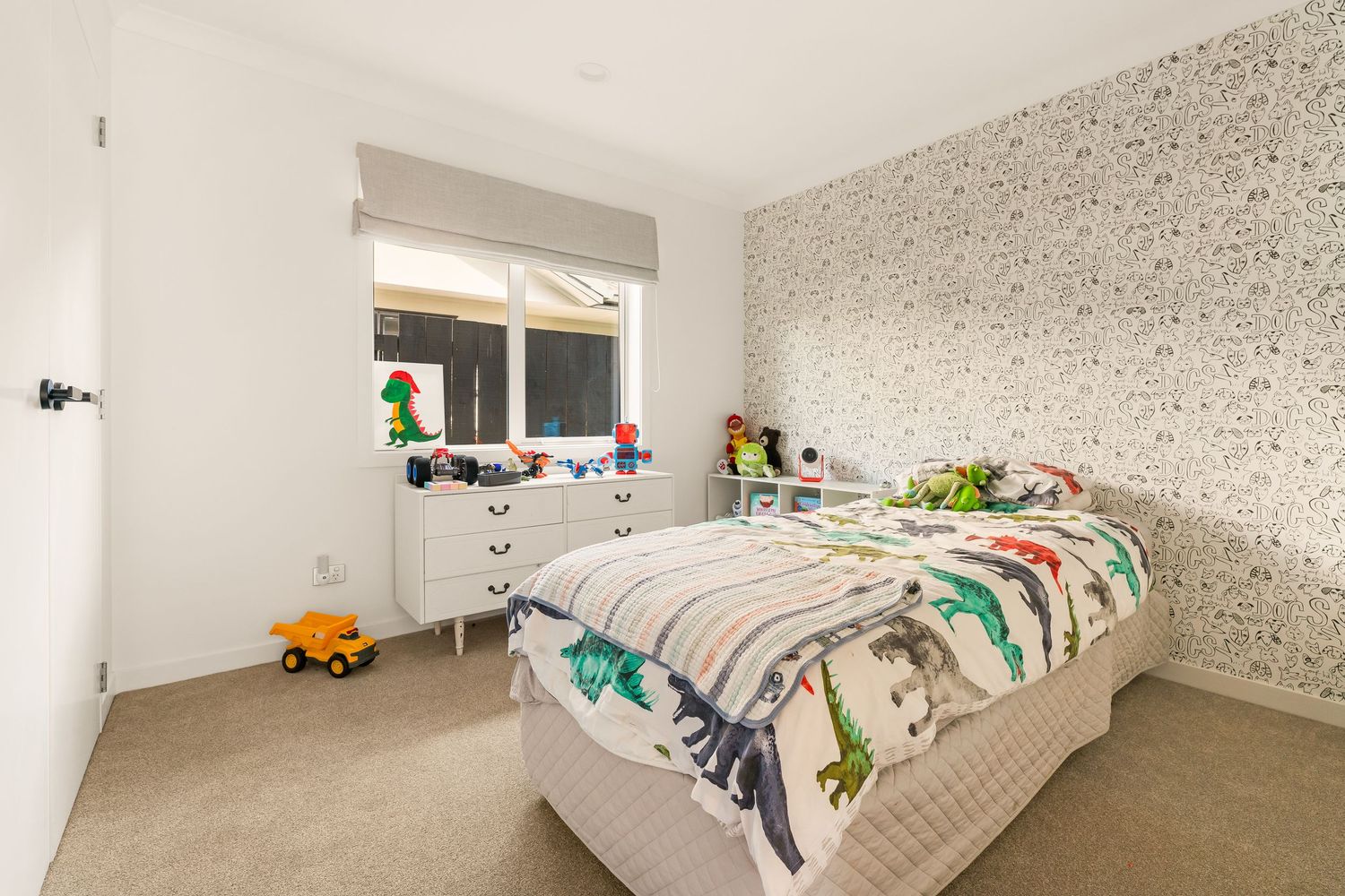 Children's bedroom with patterned wallpaper, white furniture and colorful dinosaur themed bedding.
