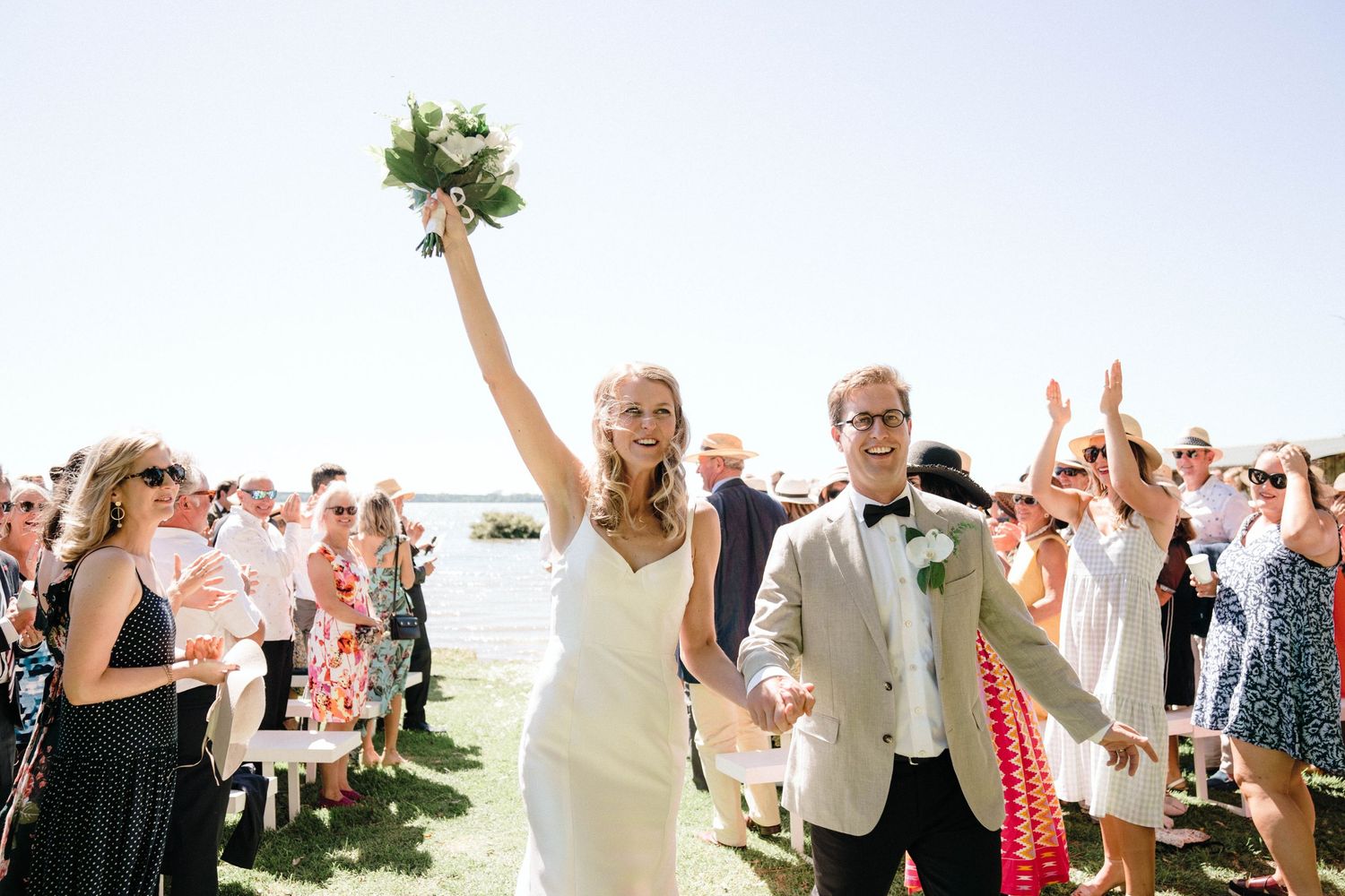 Newlyweds celebrate with raised arms and a bouquet while guests cheer during an outdoor wedding ceremony.