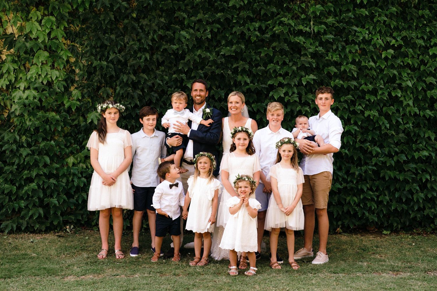 A large family group in coordinated white outfits poses together against a lush green hedge backdrop.