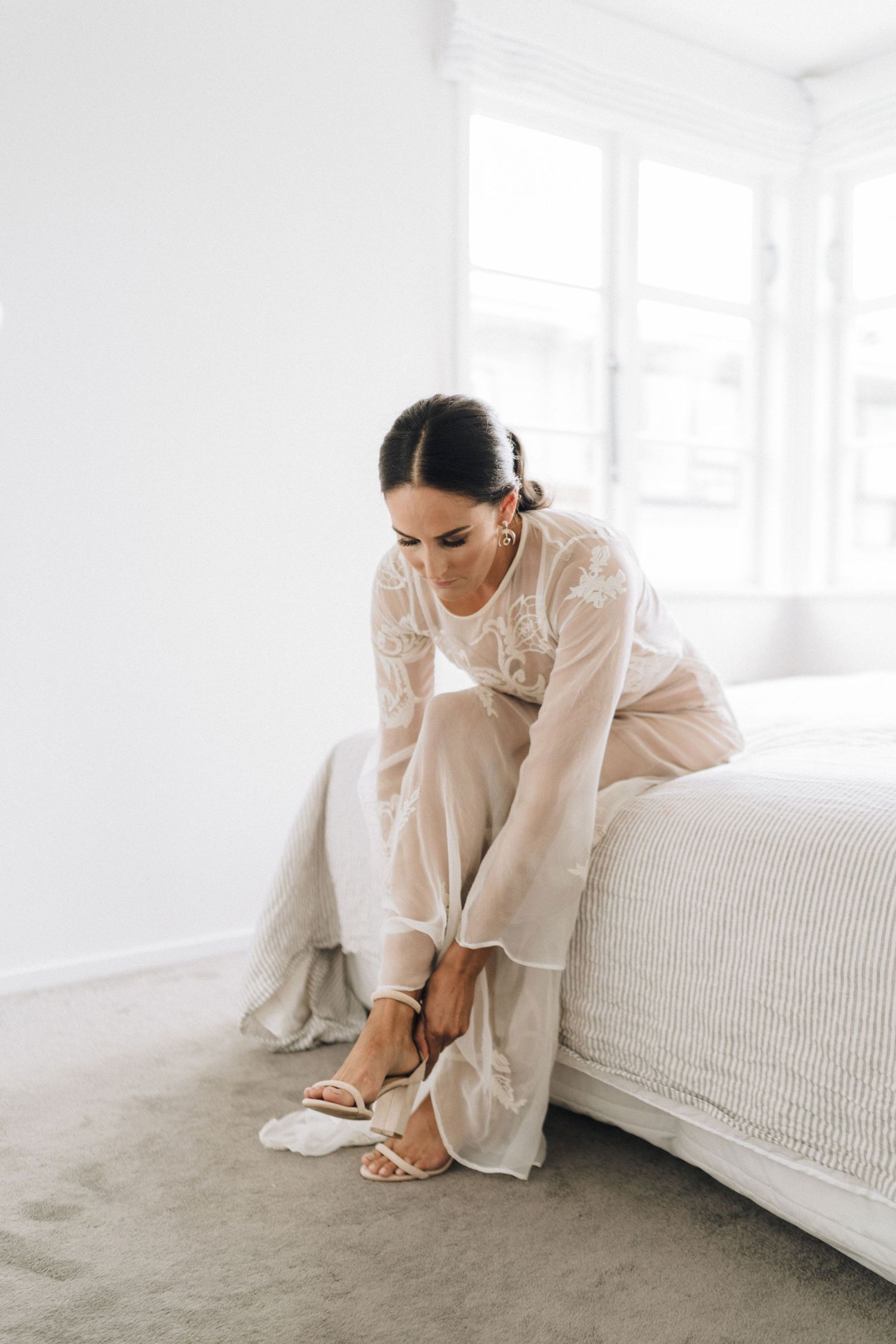 Person in white dress sitting on edge of white bed in minimalist room with natural light.
