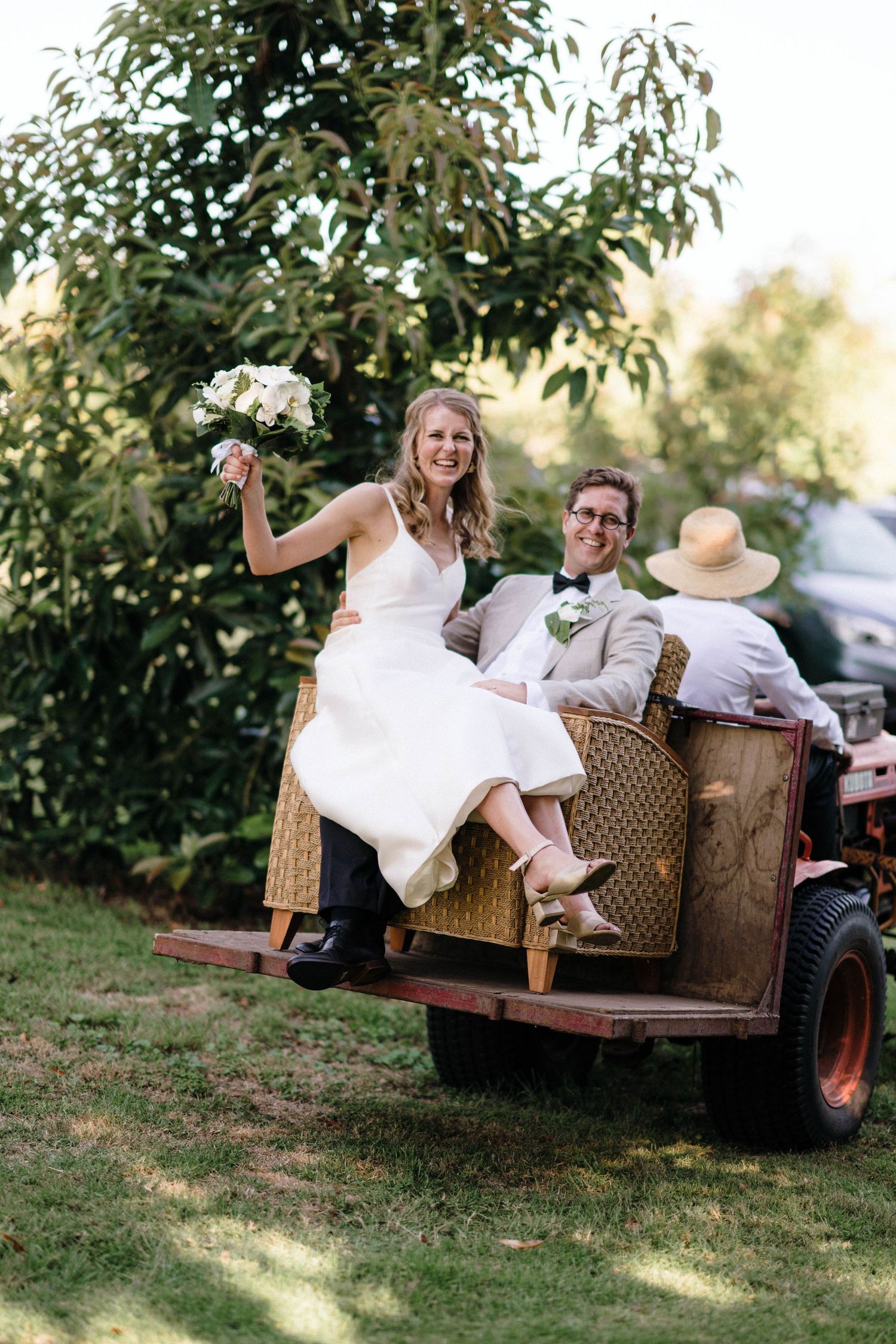 Newlyweds ride together on a rustic wicker-laden farm trailer during an outdoor countryside wedding celebration.