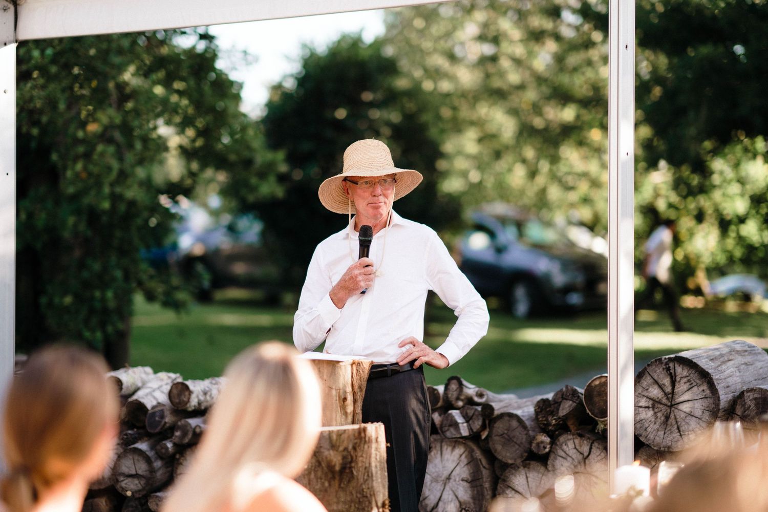 A speaker in a straw hat and white shirt addresses wedding guests at an outdoor venue.