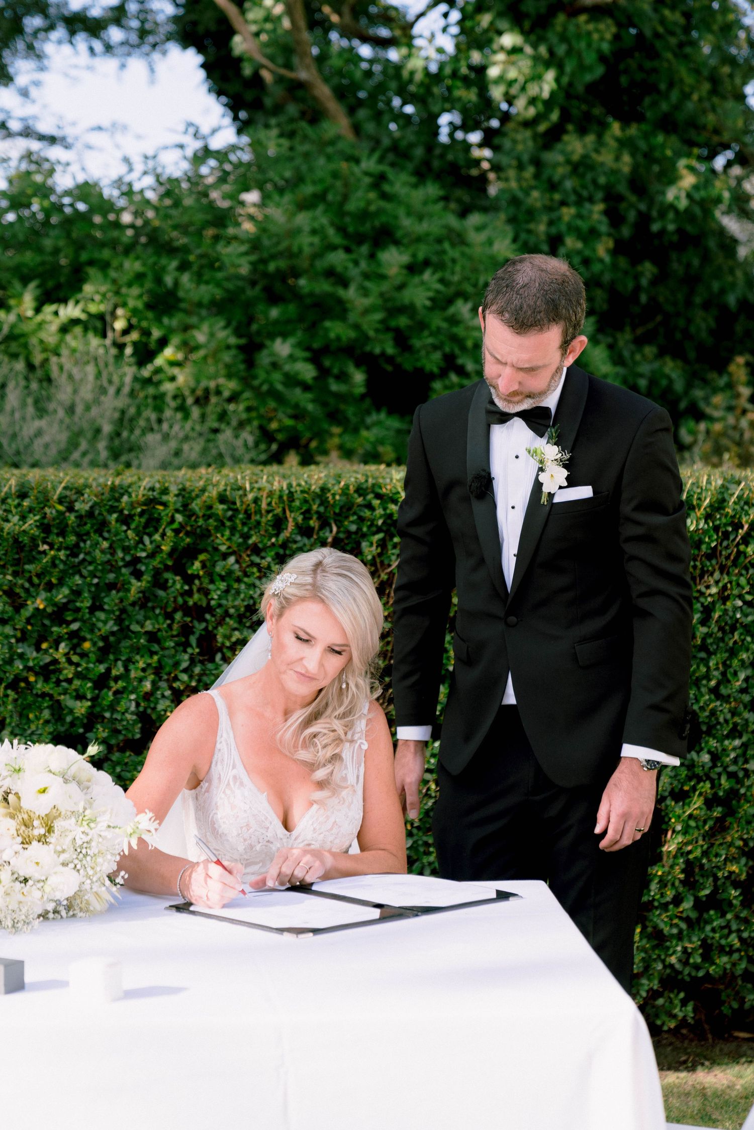 A bride in a white gown signs marriage documents at an outdoor ceremony table while the groom stands beside her.