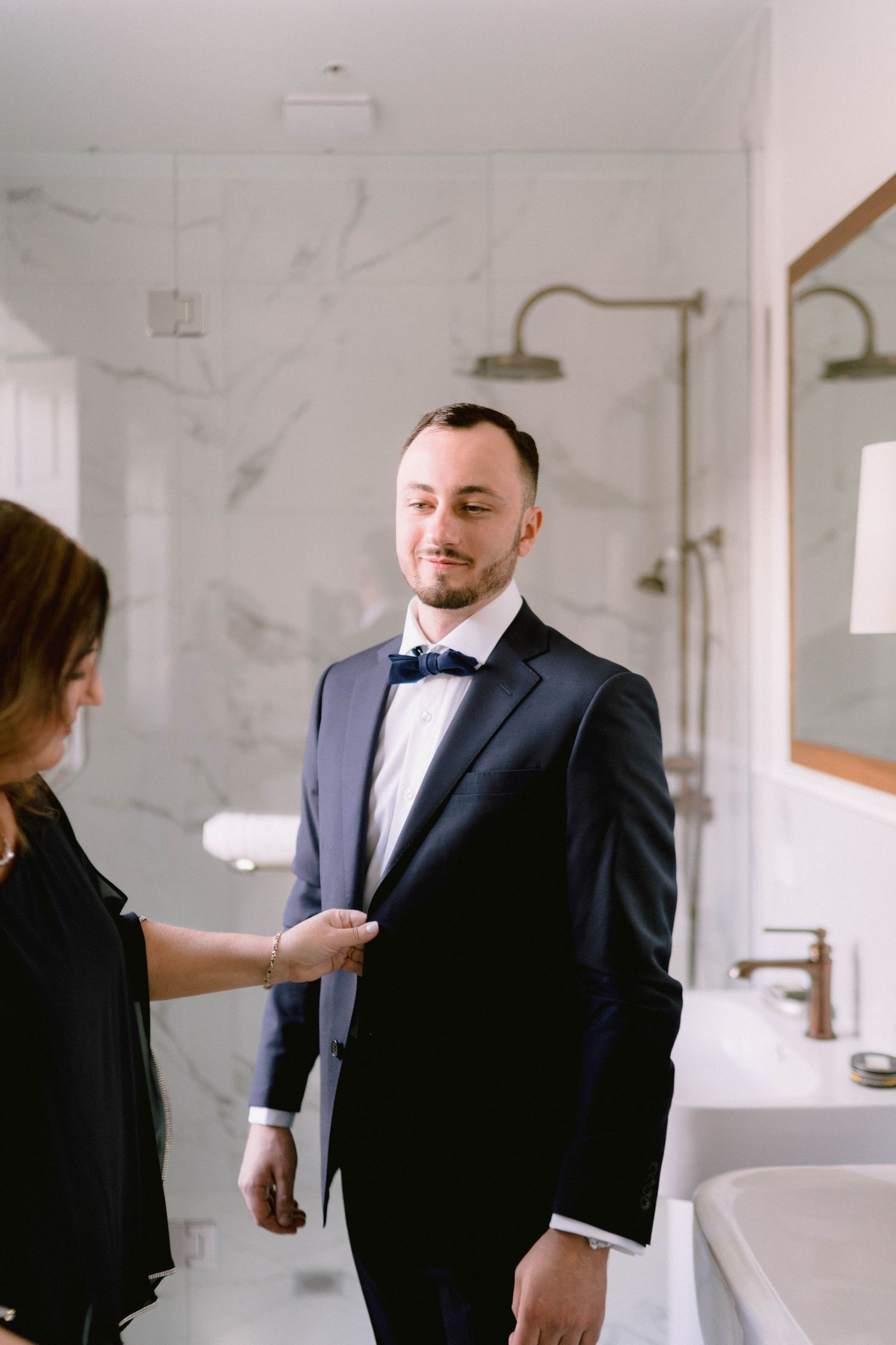 Wedding preparations showing a formal black suit being adjusted in a bright white marble room.