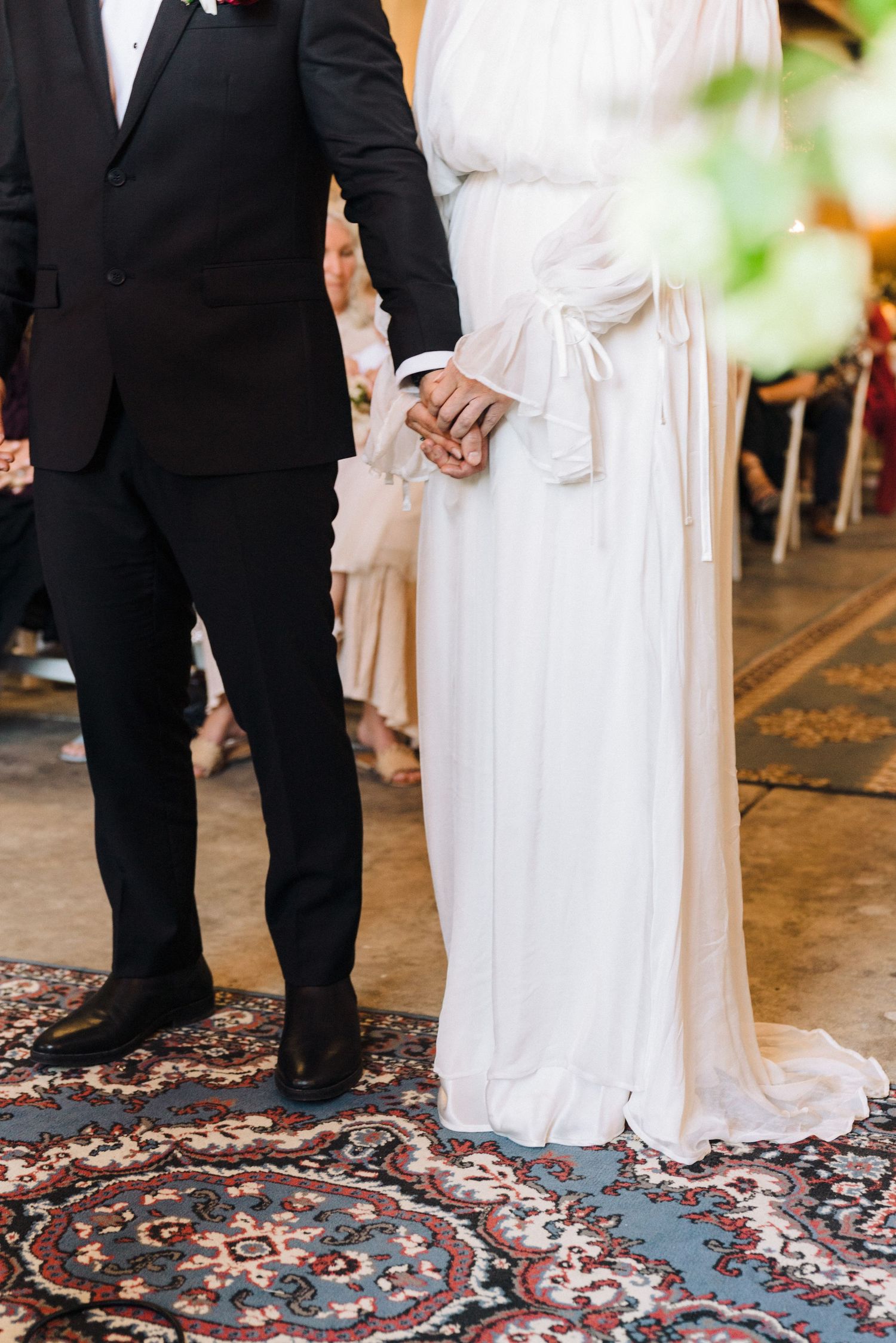 Close-up of a couple in wedding attire holding hands while standing on a patterned carpet.
