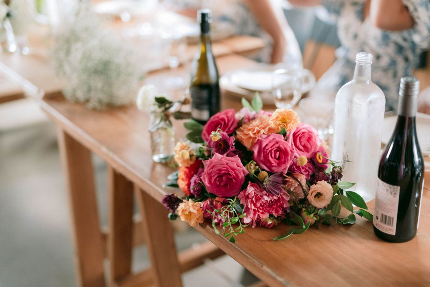Elegant floral centerpiece with pink roses and wine bottles on wooden table at wedding reception.