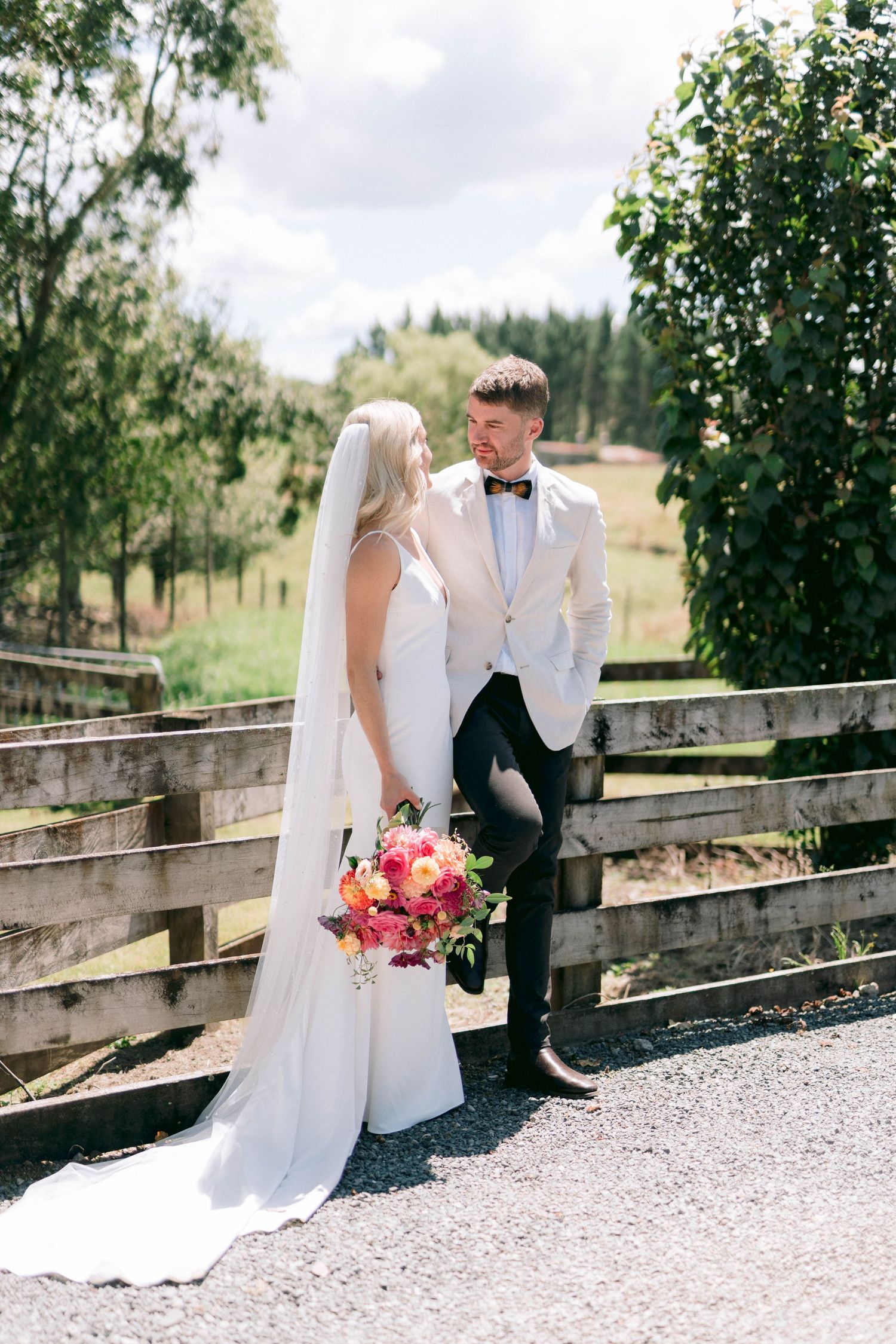 Newlyweds pose together on rustic wooden fence with colorful bouquet against countryside backdrop.