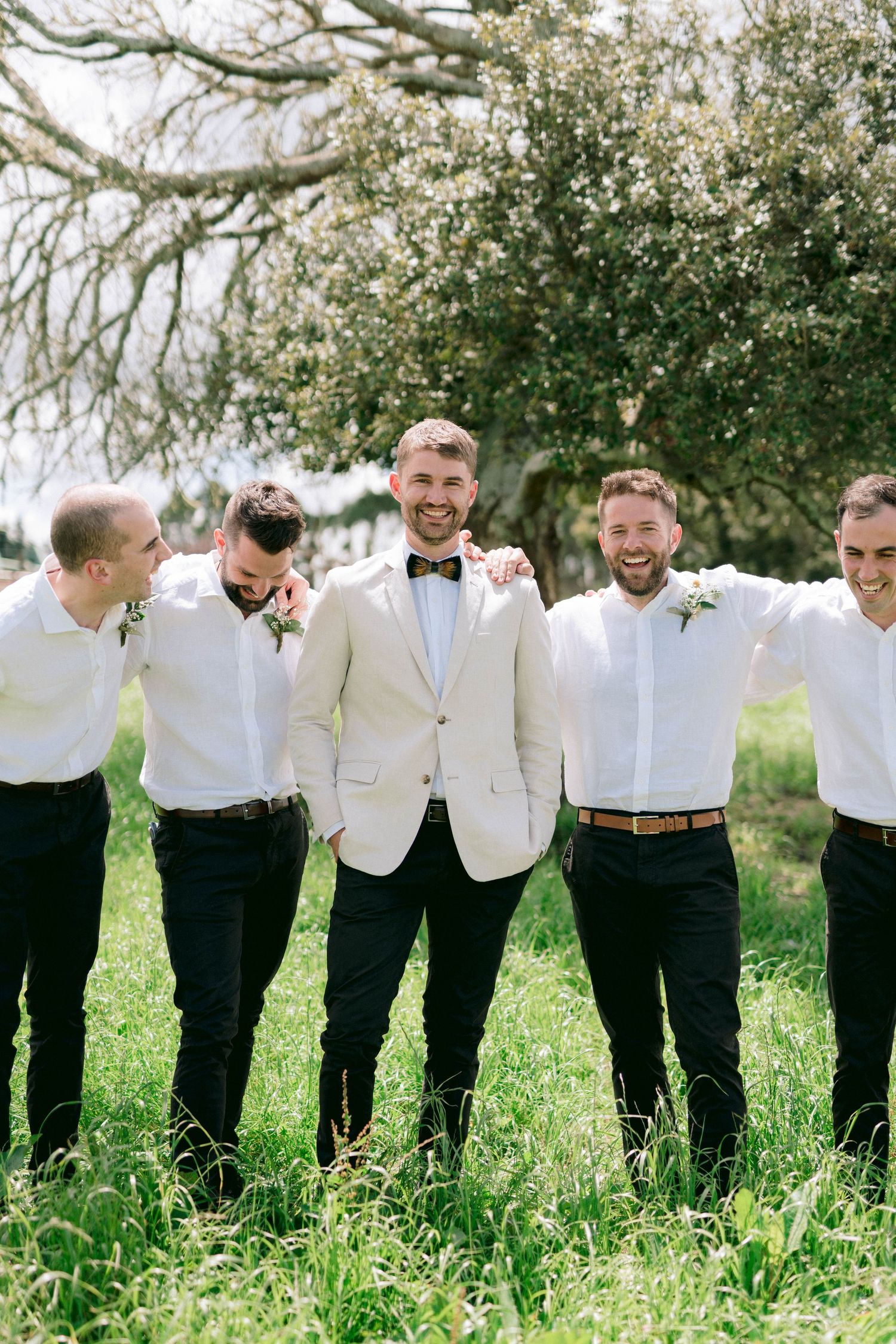 Groomsmen share a laugh while posing in matching white jackets and black pants at an outdoor wedding.