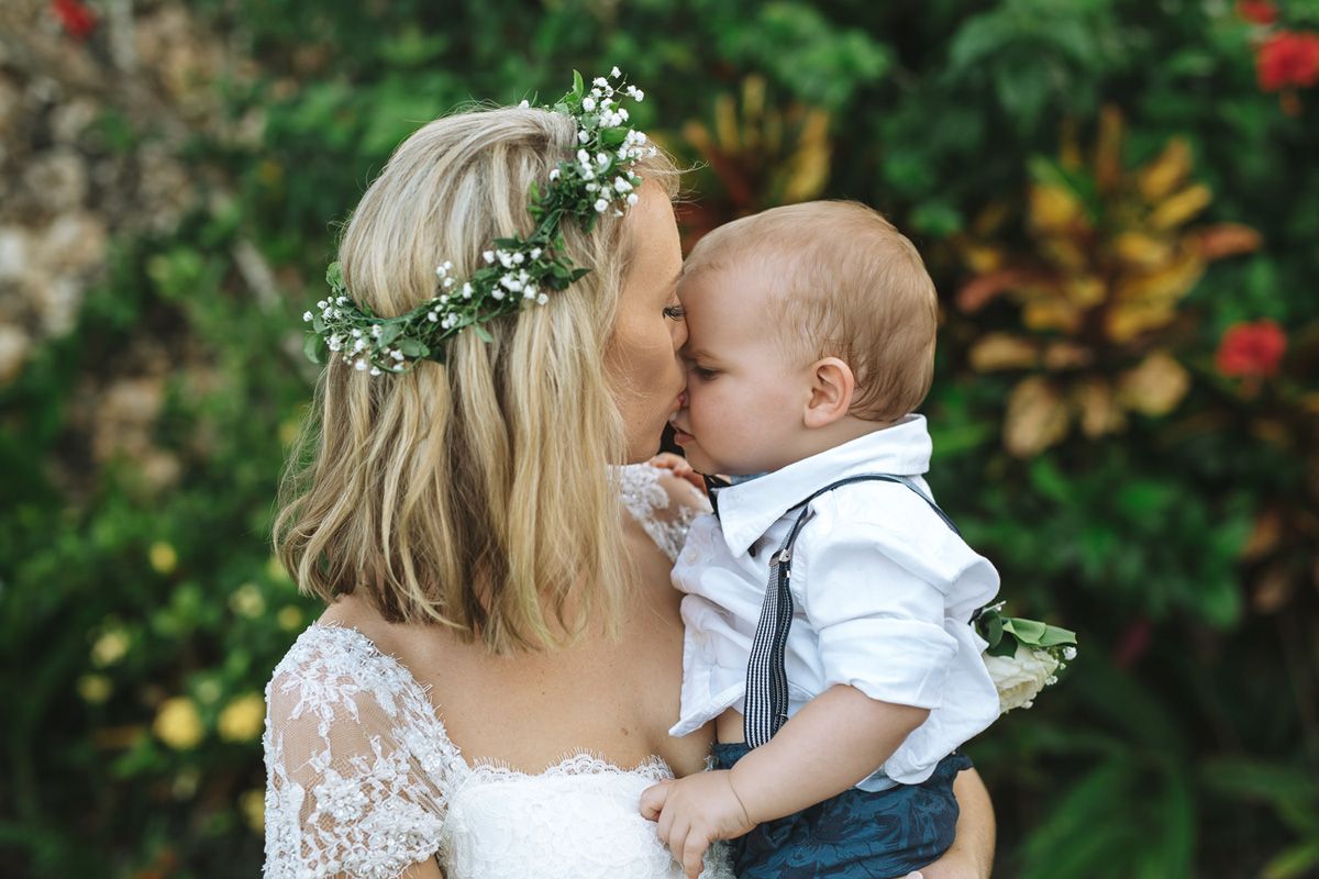 A tender moment between a bride wearing a floral crown and a baby in a white shirt against a garden backdrop.