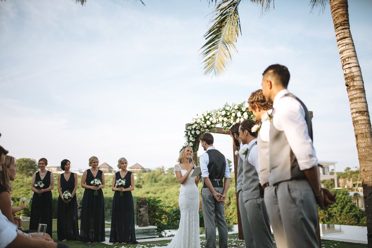 An outdoor wedding ceremony takes place under palm trees with bridesmaids in dark dresses and groomsmen in grey suits.