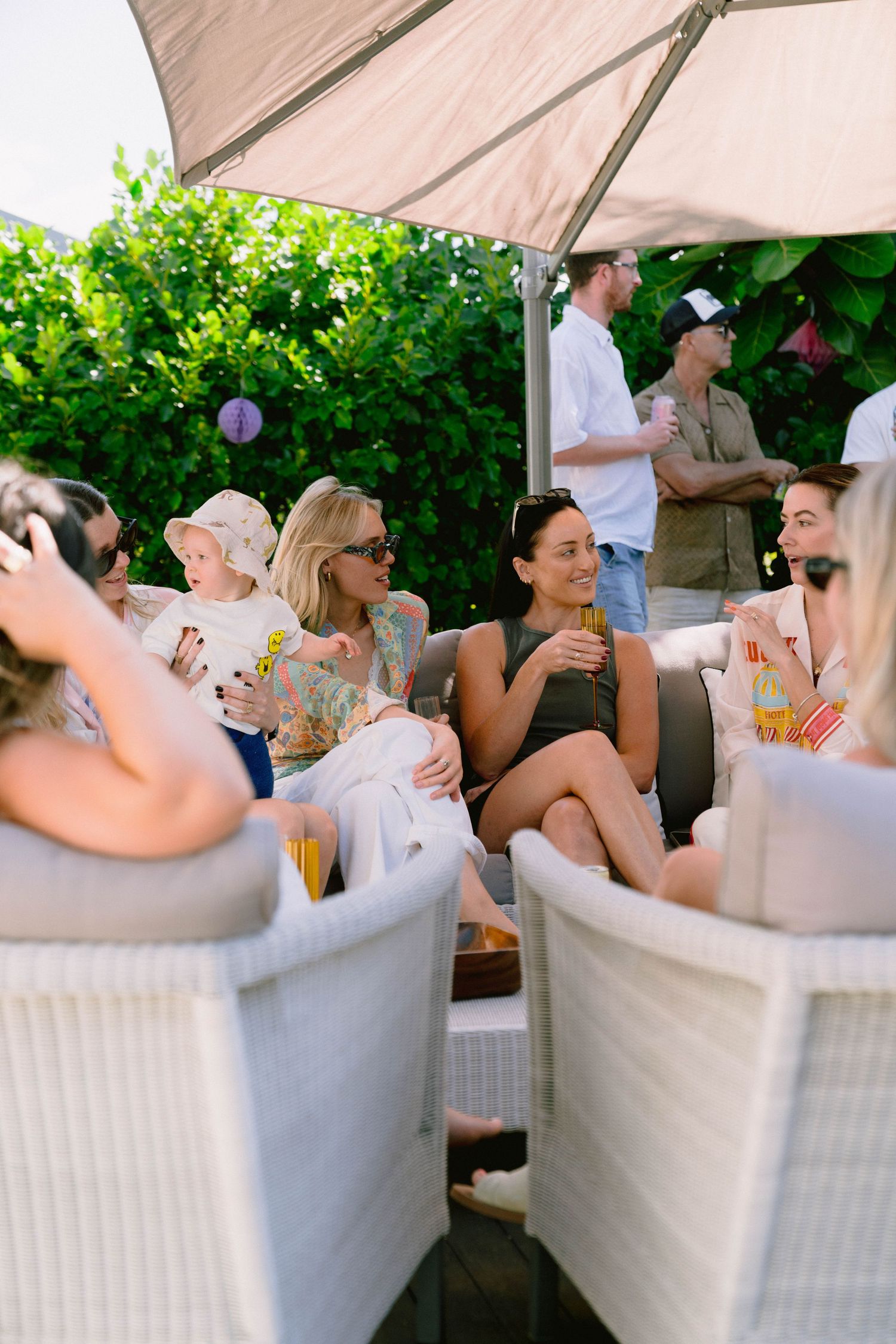 A group relaxes on comfortable white wicker outdoor furniture under a beige umbrella at a social gathering.