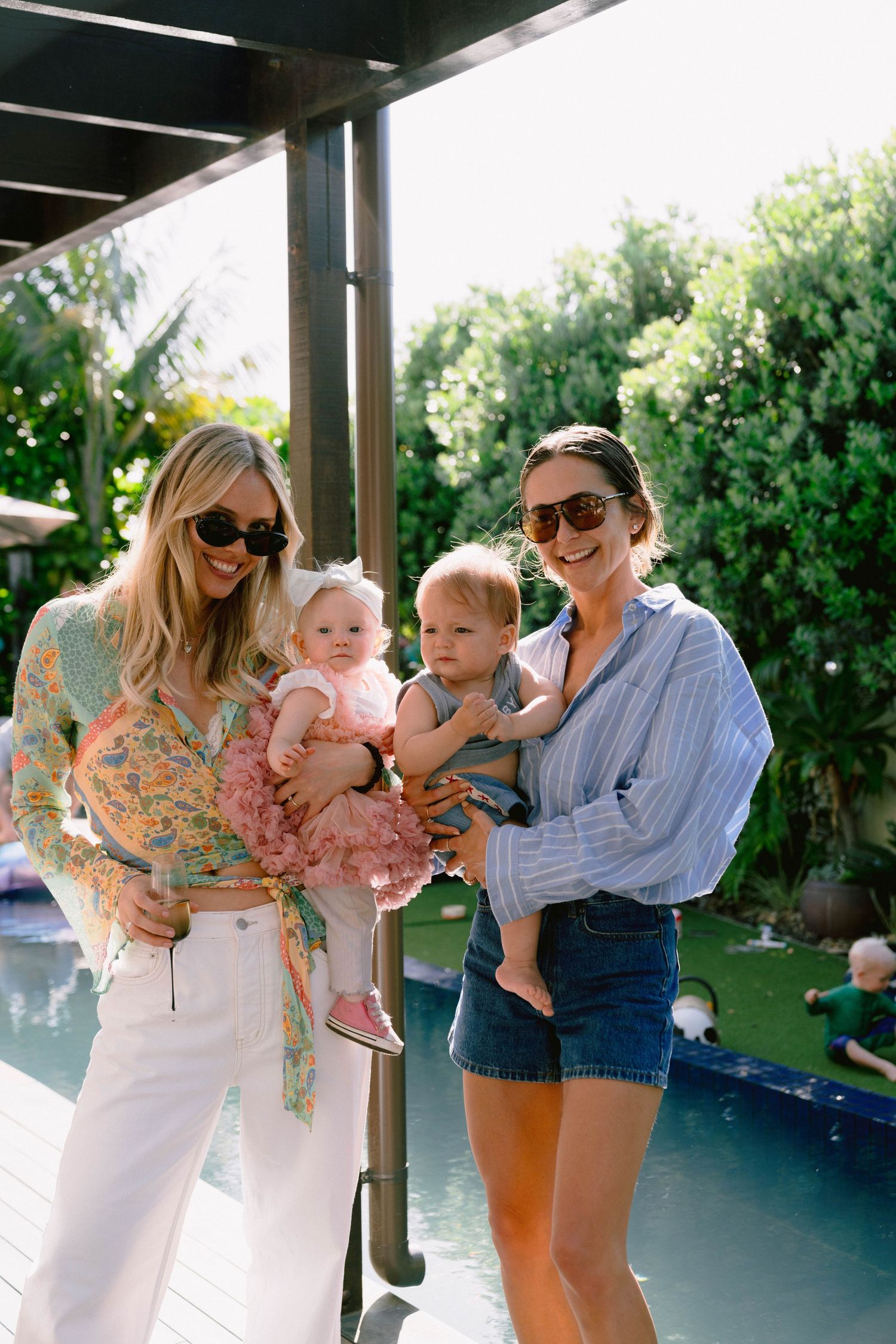 Two people holding babies next to a swimming pool on a sunny day.