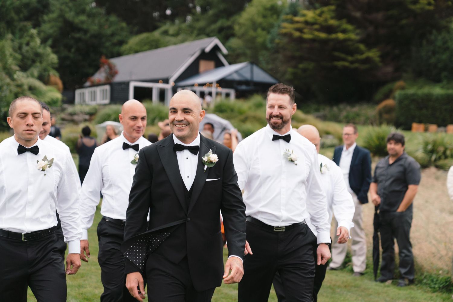 Groomsmen in black tuxedos and bow ties walk together smiling on grounds at outdoor wedding venue.
