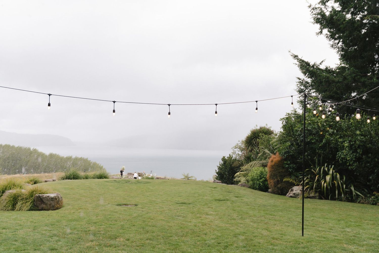 String lights hang over a grassy hilltop overlooking a misty lake or ocean view on an overcast day.