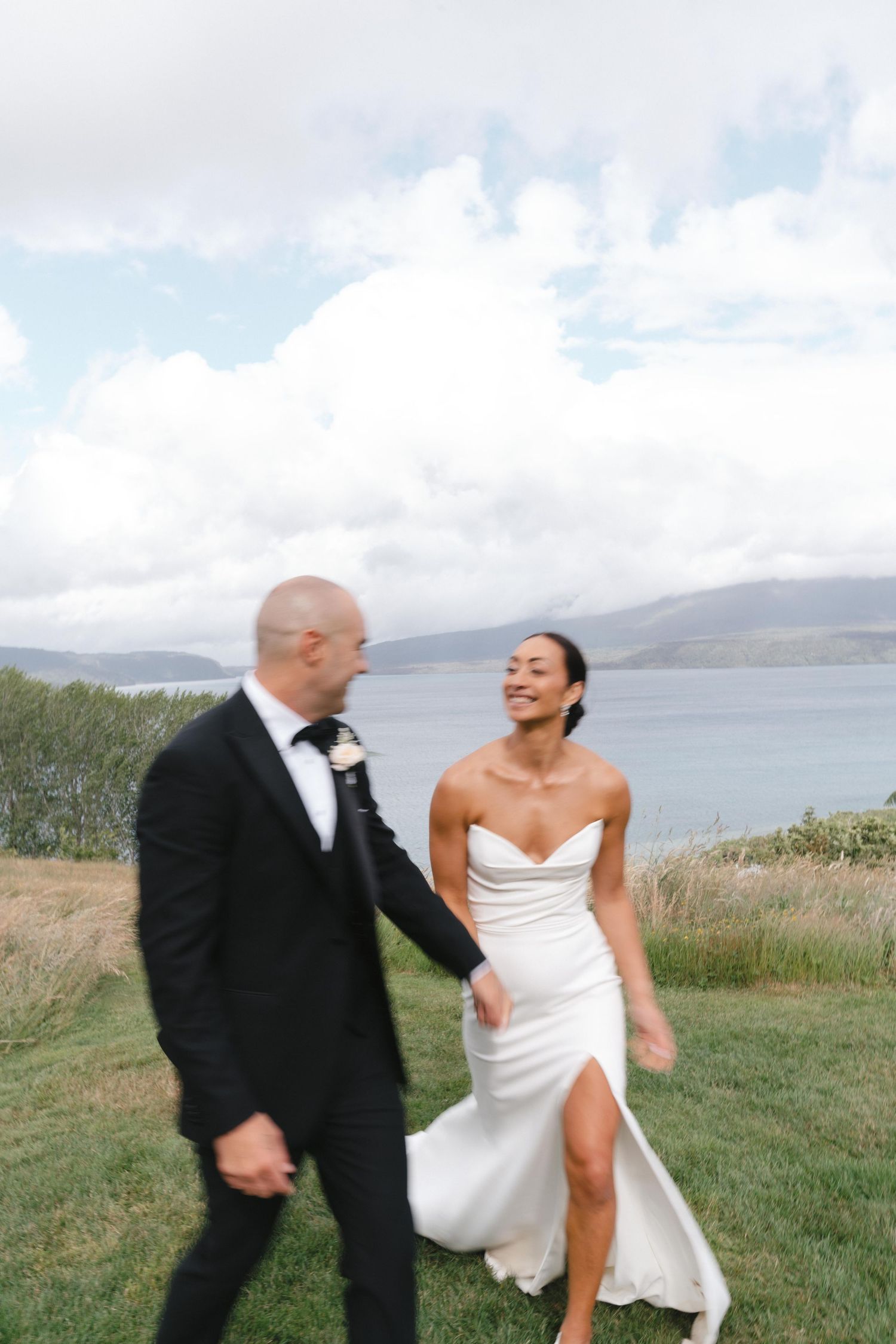 Newlyweds share playful moment on scenic coastal overlook with water views.