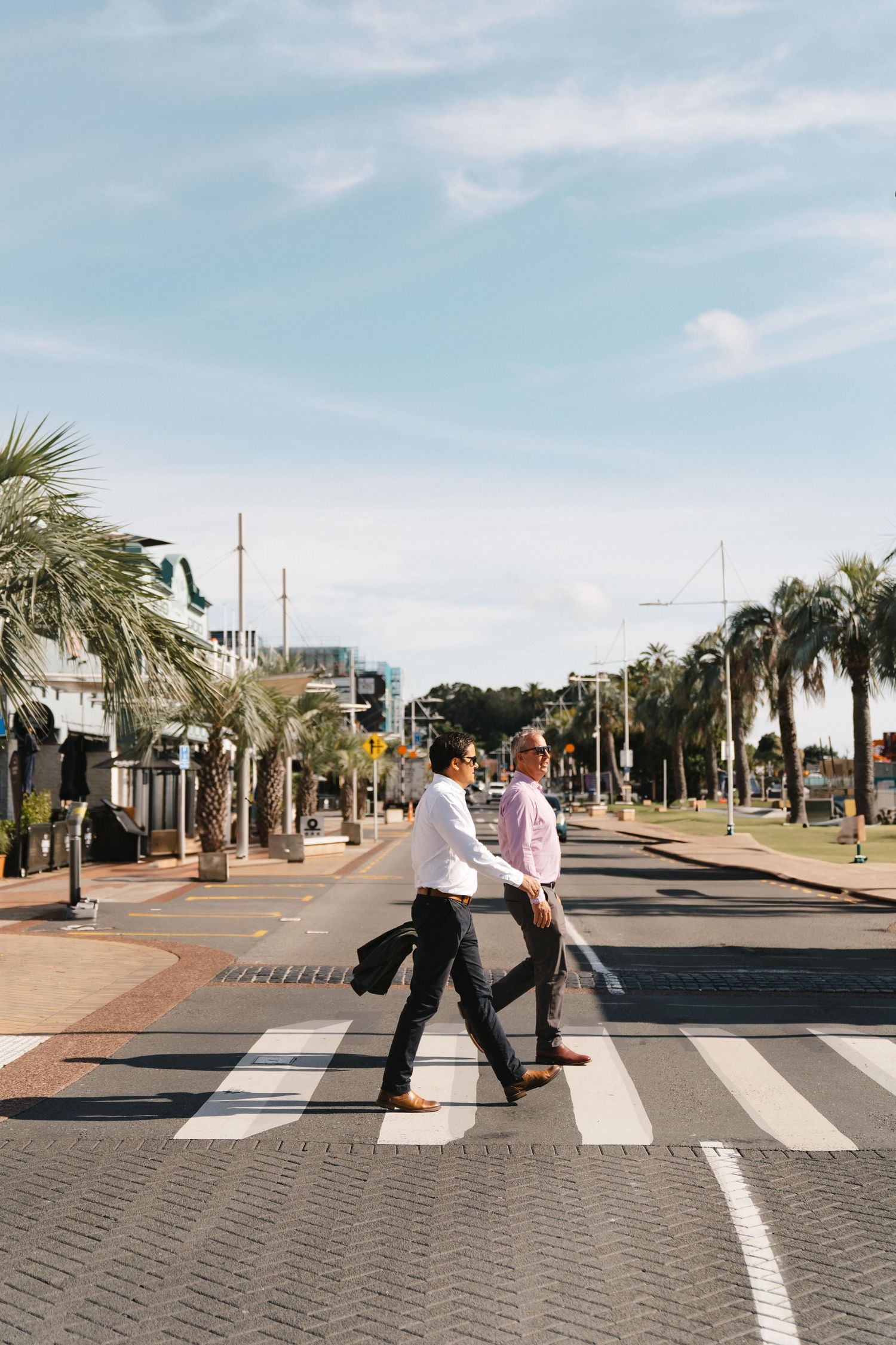 People walking across a palm tree-lined crosswalk on a sunny street.
