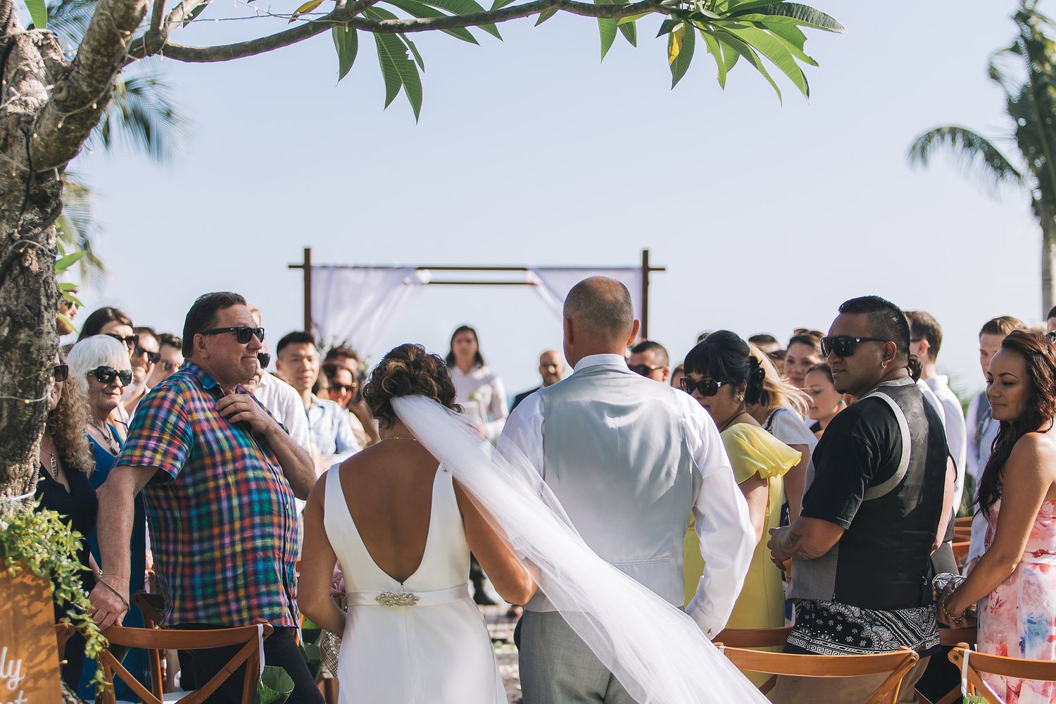 Outdoor beach wedding ceremony with guests gathered around a white fabric-draped arch under palm trees.