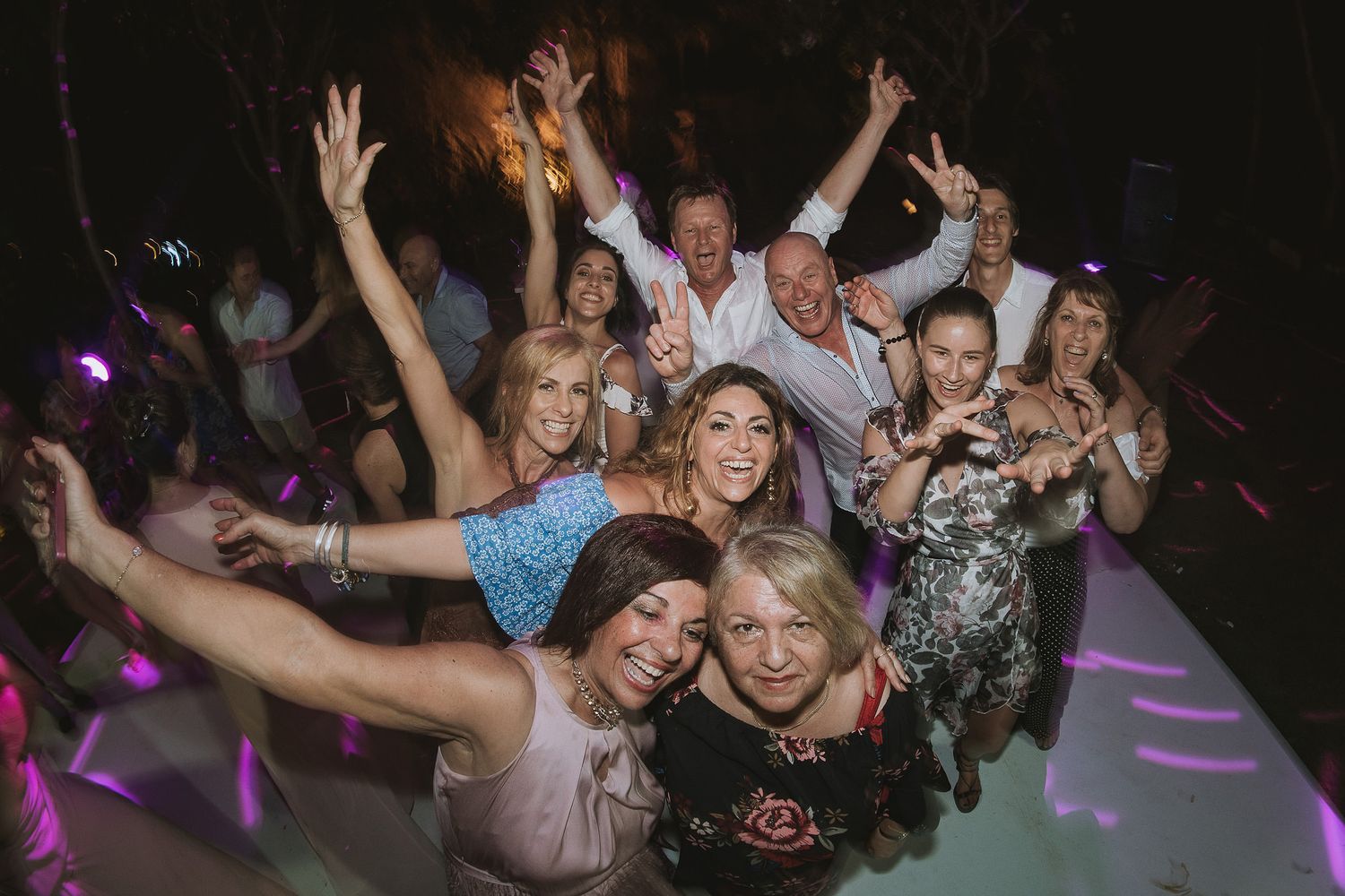 Wedding guests celebrate on the dance floor with energetic poses under purple lighting.