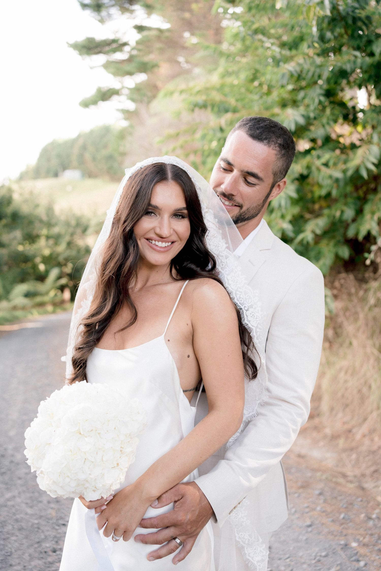 Intimate wedding portrait of couple in white embracing on a forest path.