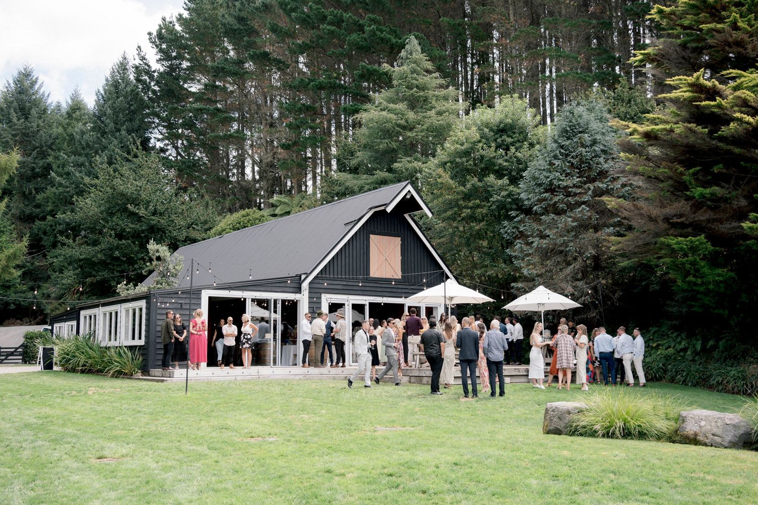 Black barn-style event venue with guests gathered outside under tall pine trees.