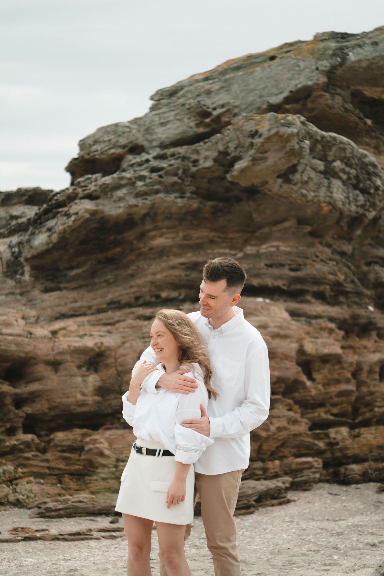 A romantic couple embraces against a backdrop of weathered coastal cliffs wearing coordinated white outfits.