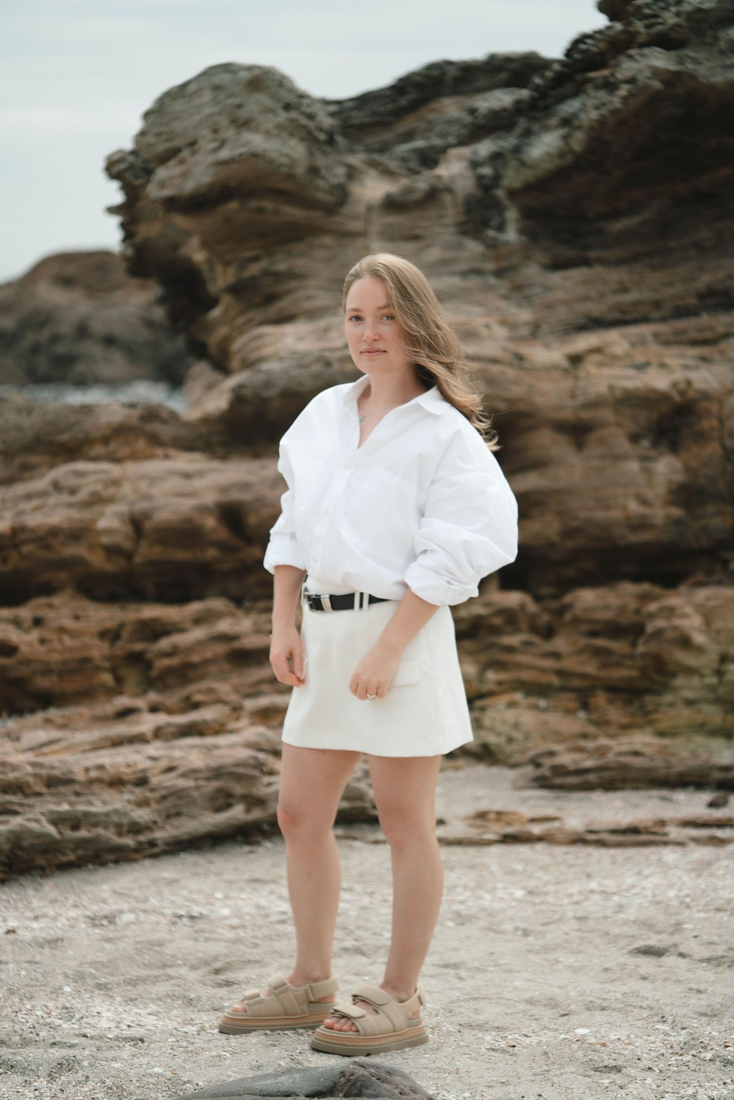 Person in white button-down shirt and mini skirt poses on rocky beach landscape with rugged cliffs in background.