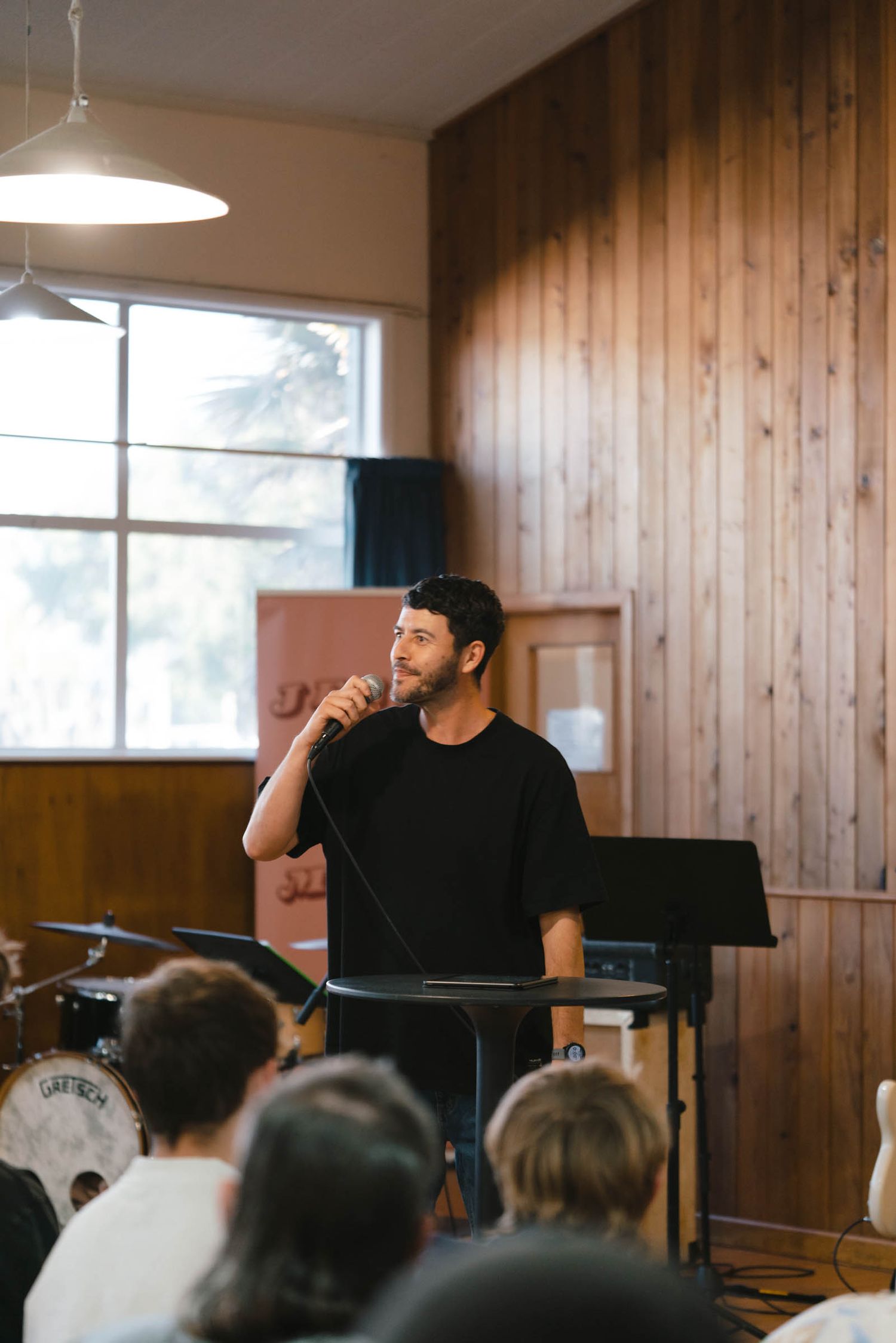 Musician stands at a music stand in front of an audience in a wood-paneled room with natural lighting.
