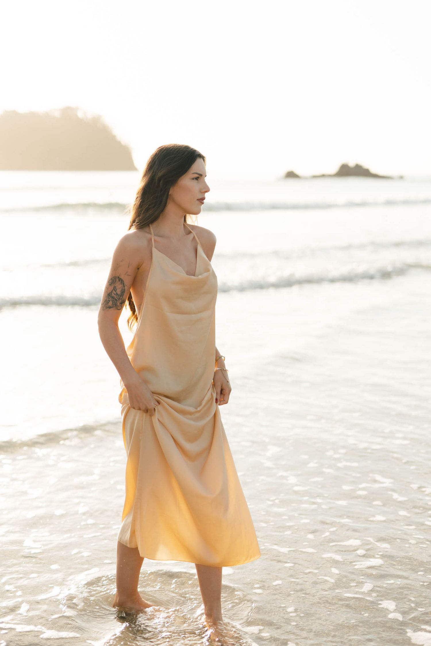 Elegant silk dress catches golden light as waves lap at shoreline during sunset.