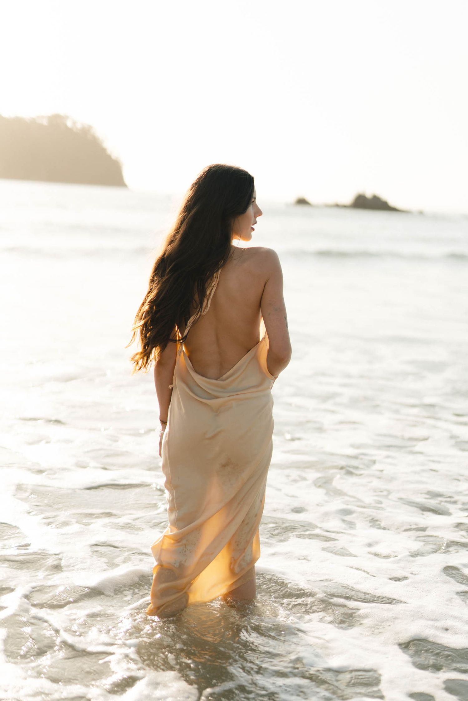 Silhouette wades through golden sunset waves in flowing dress.