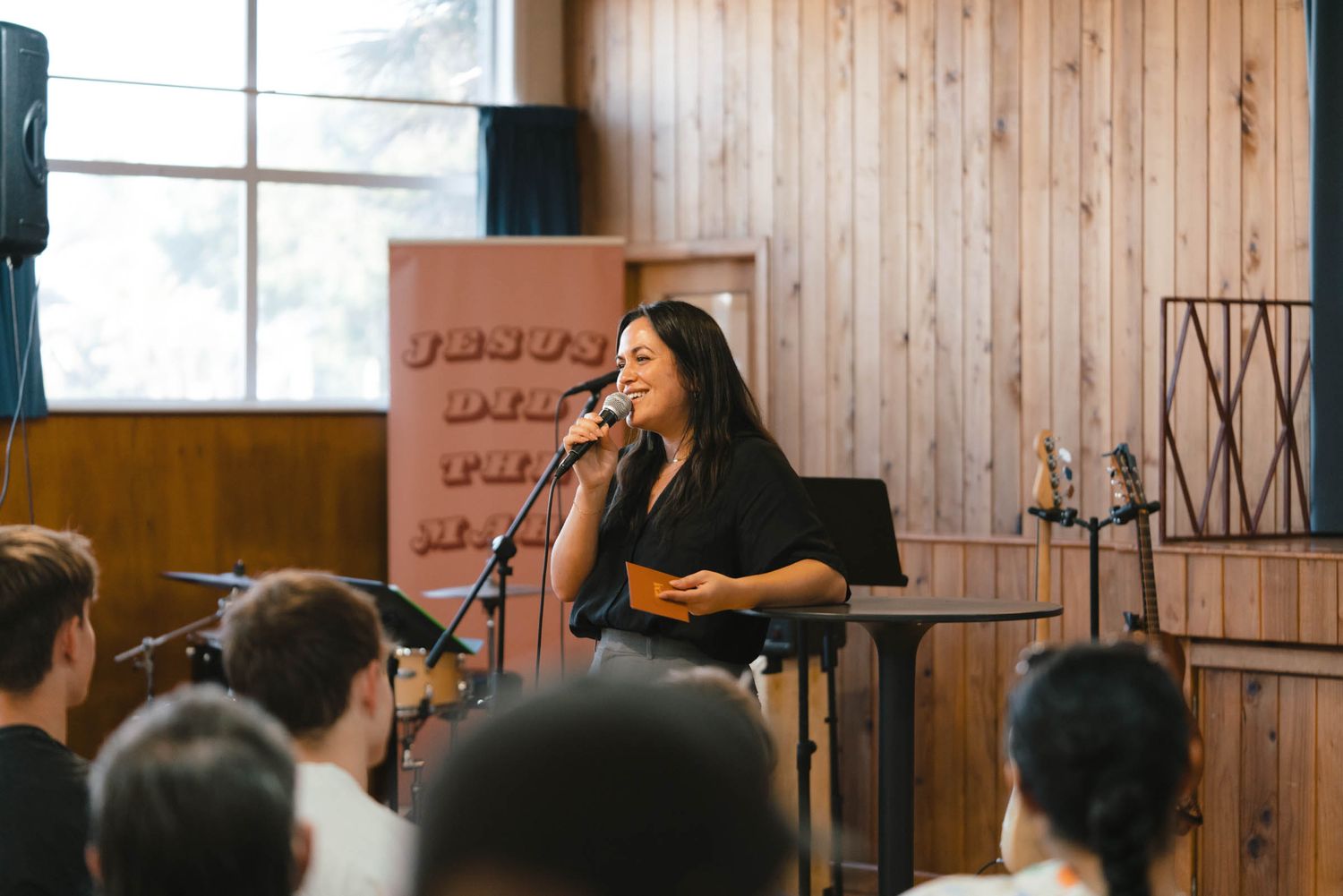 A person performs on stage with a microphone in front of an audience in a wood-paneled room.