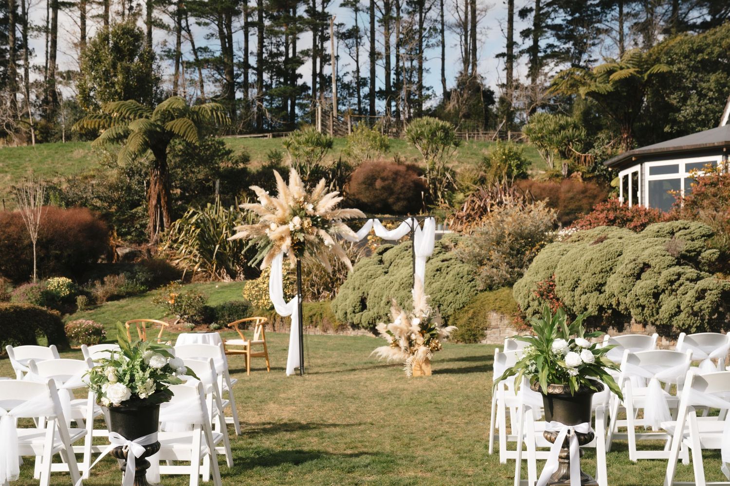 A rustic outdoor wedding ceremony setup with white chairs and pampas grass arch decorations on a grassy hillside.