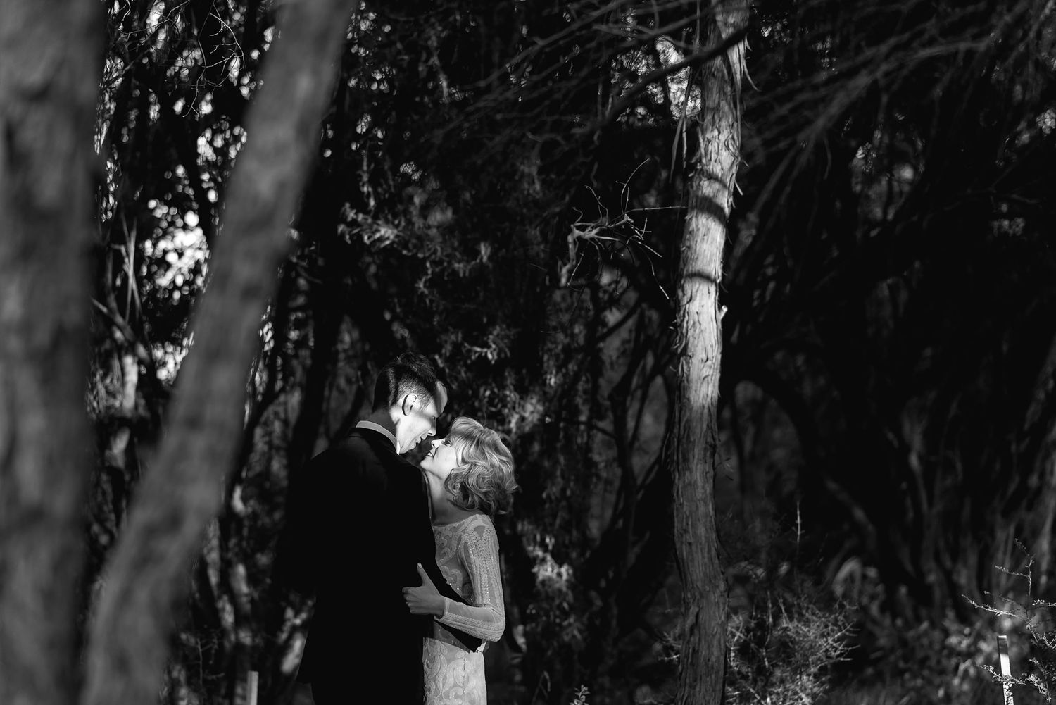 A black and white photo captures a romantic moment between a couple embracing in a wooded forest setting.