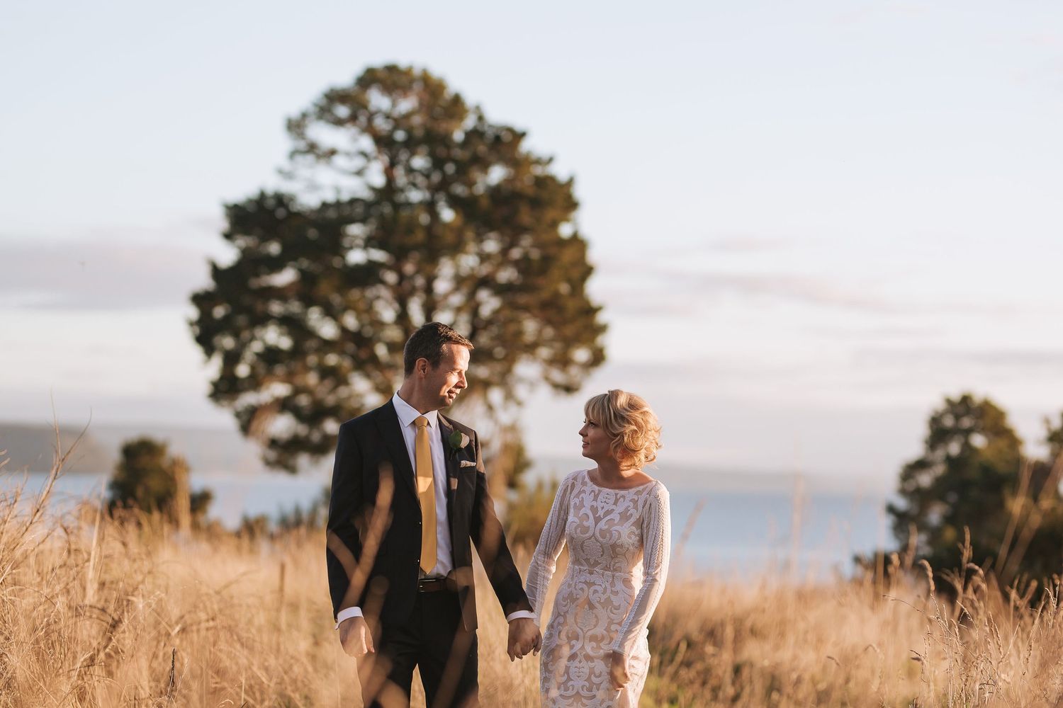 A couple walks hand in hand through a golden field at sunset with a tall pine tree and ocean view in the background.