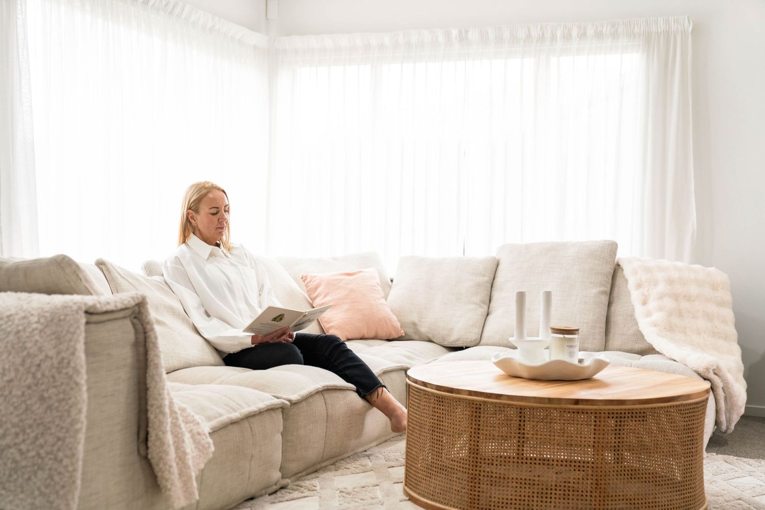 Person relaxing in cozy living room on beige couch with round coffee table and sheer white curtains.
