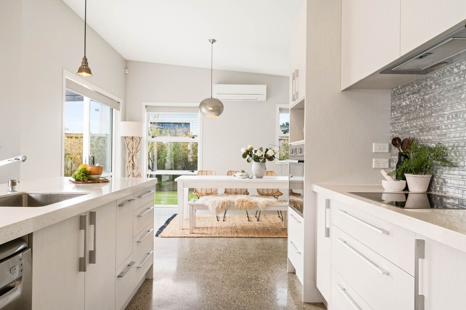 Modern white kitchen with polished concrete floors opens to bright dining area featuring pendant lights and garden views.