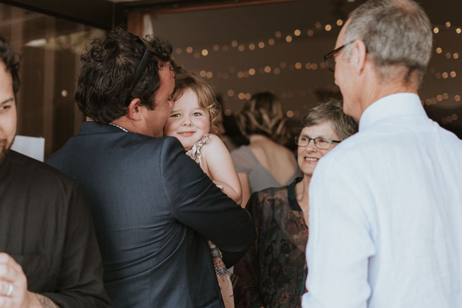 A joyful gathering at an indoor event with string lights overhead as people interact warmly in a casual atmosphere.