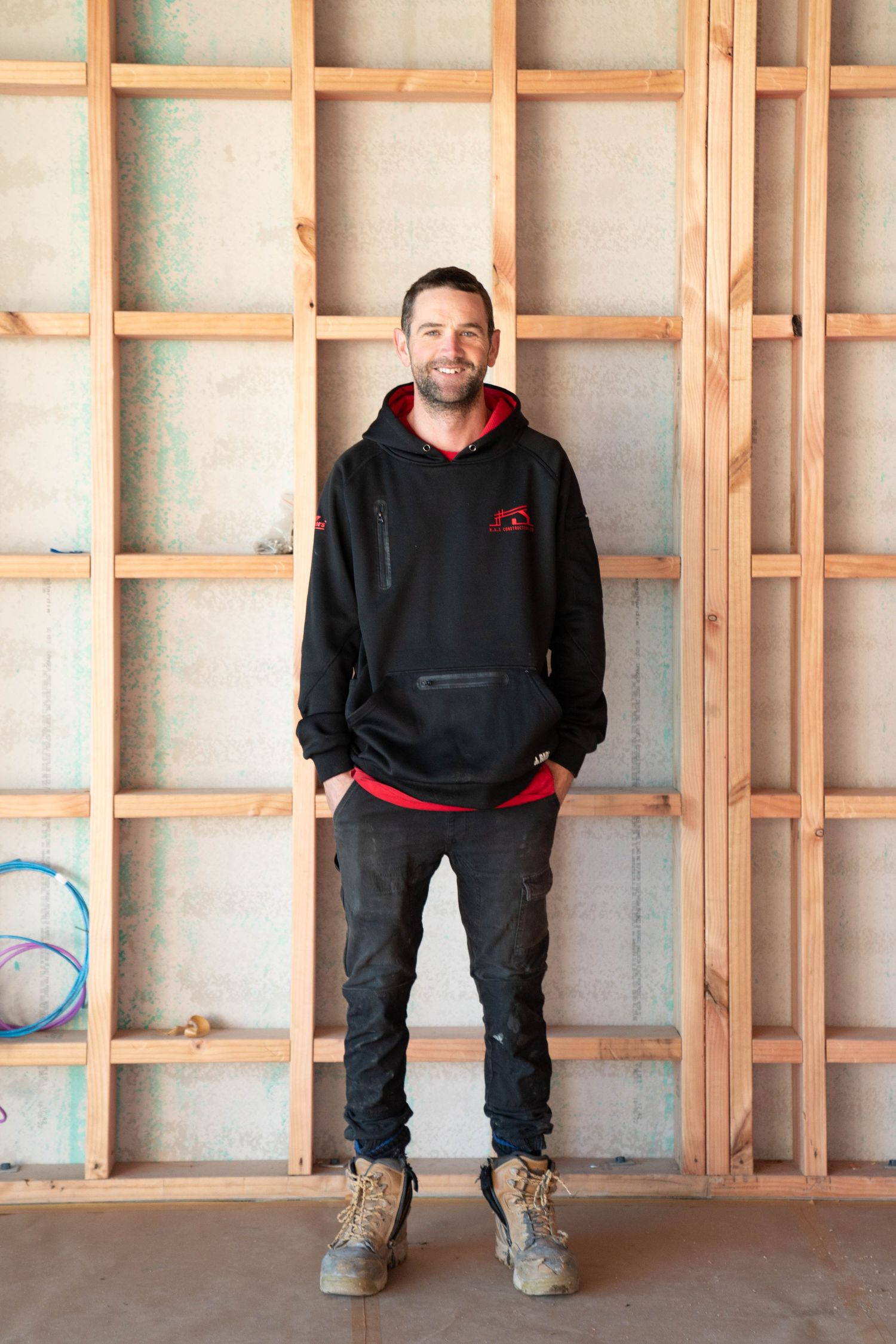 Person wearing black hoodie and pants sits casually on wooden framing in unfinished construction or renovation space.