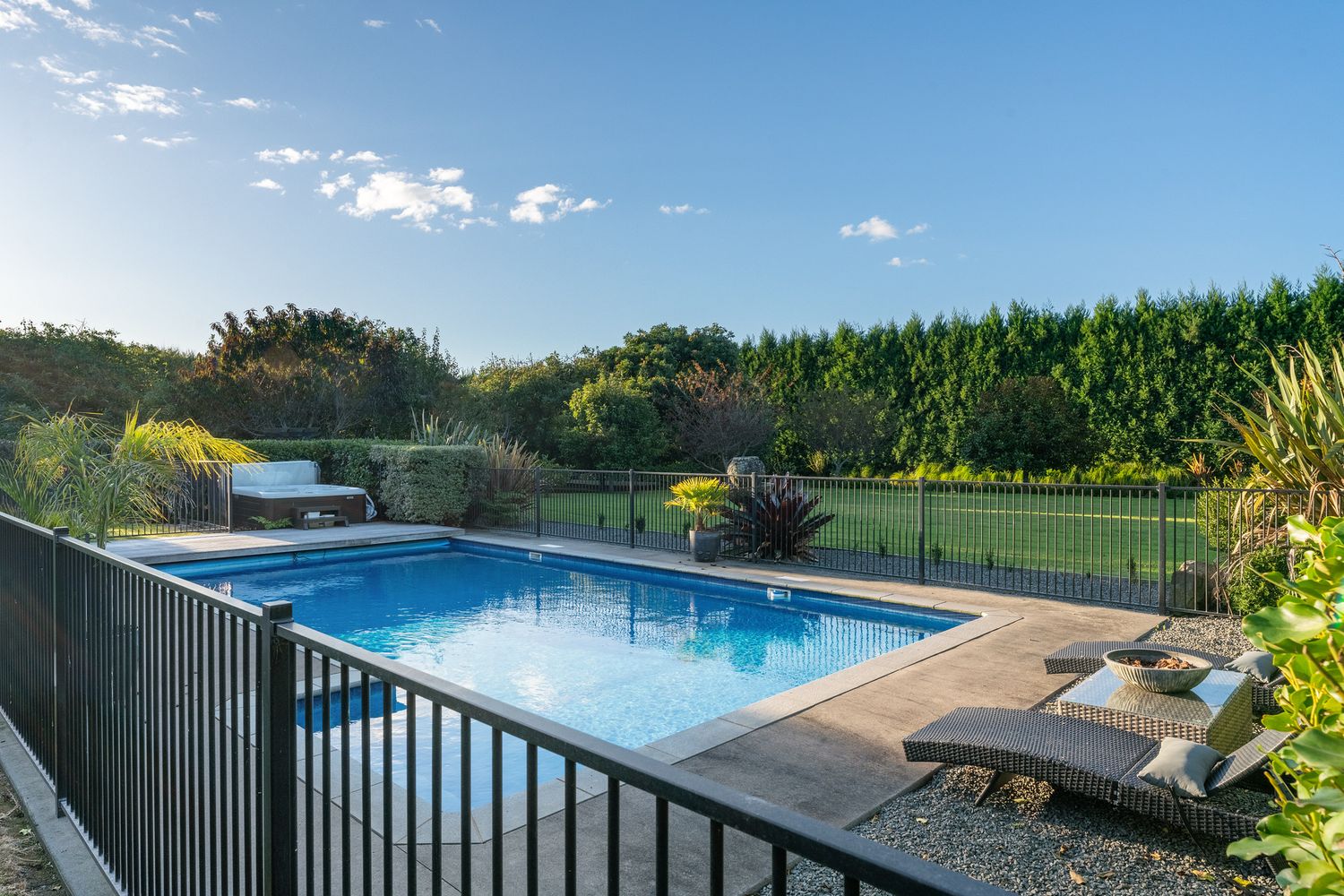 Rectangular inground pool with loungers surrounded by black fencing and lush landscaping under blue skies.